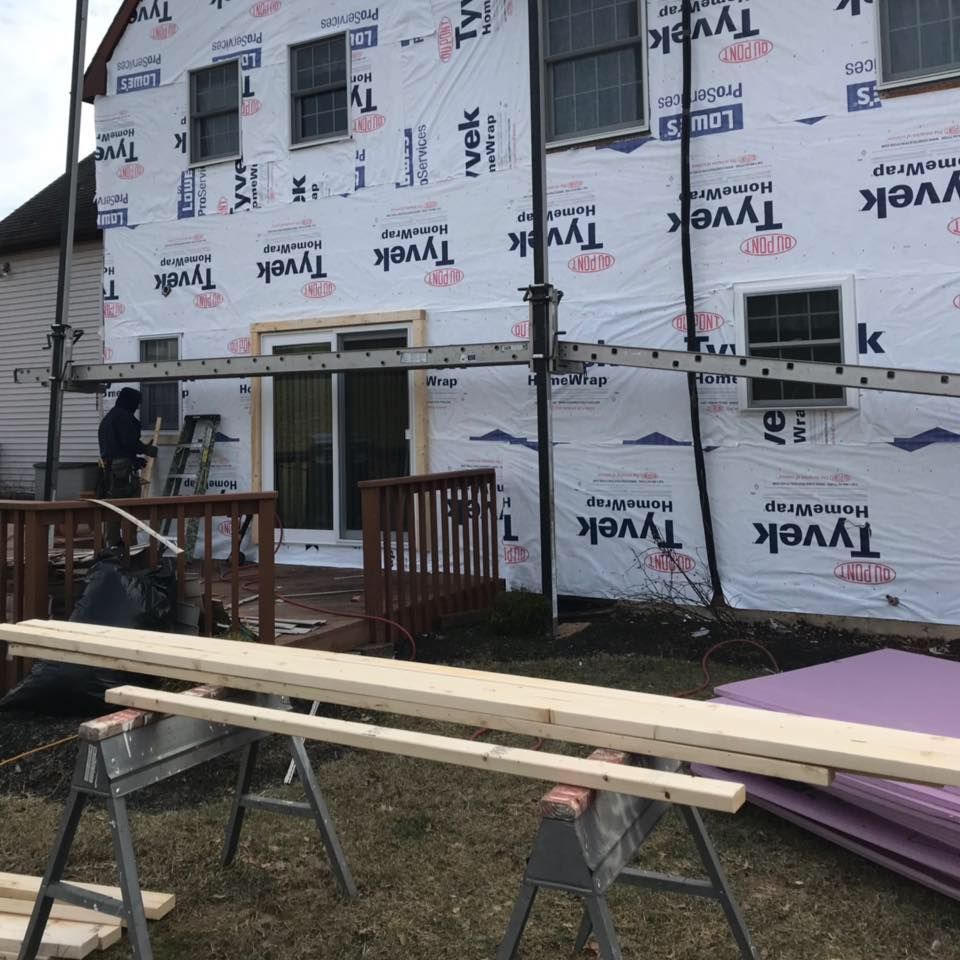 House under construction, covered with Tyvek. A worker stands on scaffolding near a sliding door, lumber on sawhorses.
