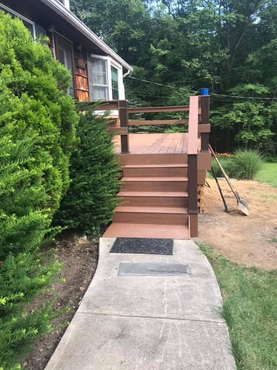 Brown deck with stairs leading to a house, concrete path, green shrubbery on the left, and trees in the background.