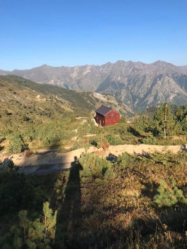 Red cabin nestled in a valley among mountains, under a clear blue sky. Shadow of person in foreground.