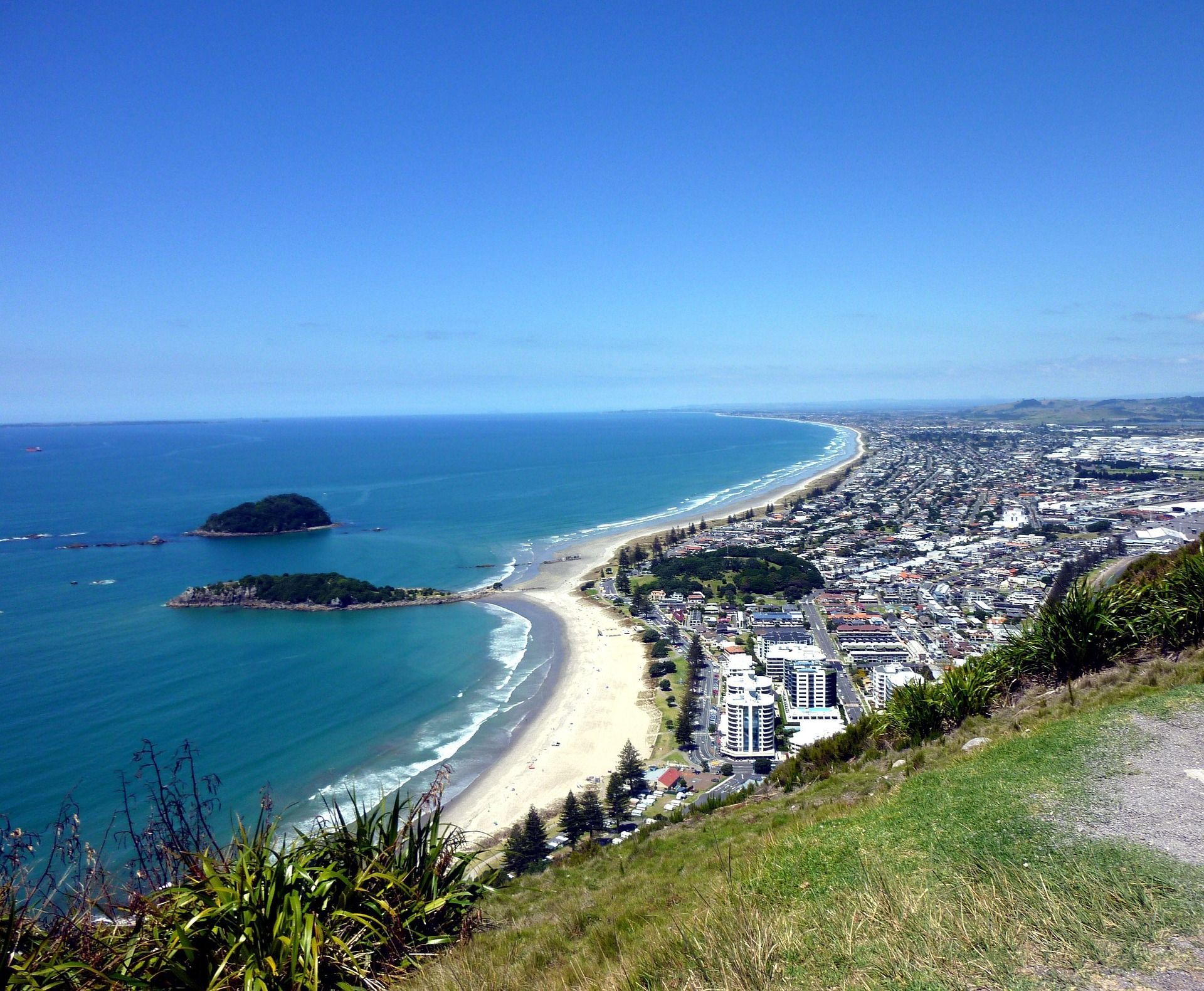 Coastal view: sandy beach, turquoise water, island, city buildings under a bright blue sky.