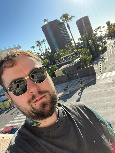 Selfie of a person wearing sunglasses on a sunny city rooftop with palm trees and tall buildings behind them