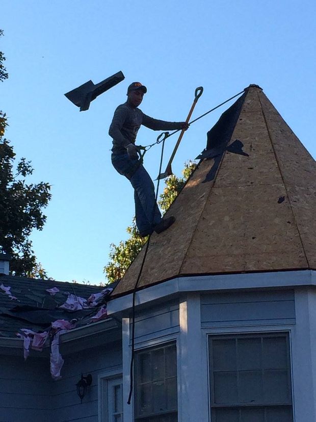 Roofer on a house roof, removing shingles, attached to safety rope; blue sky background.