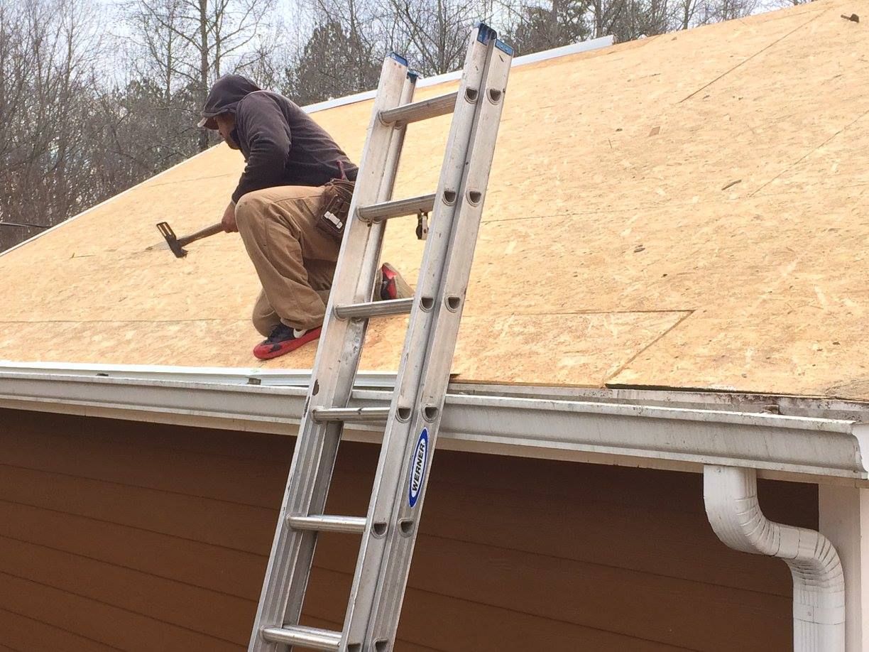 Person hammering on a roof, standing on a ladder. Brown pants, dark jacket. Gutters and house visible.