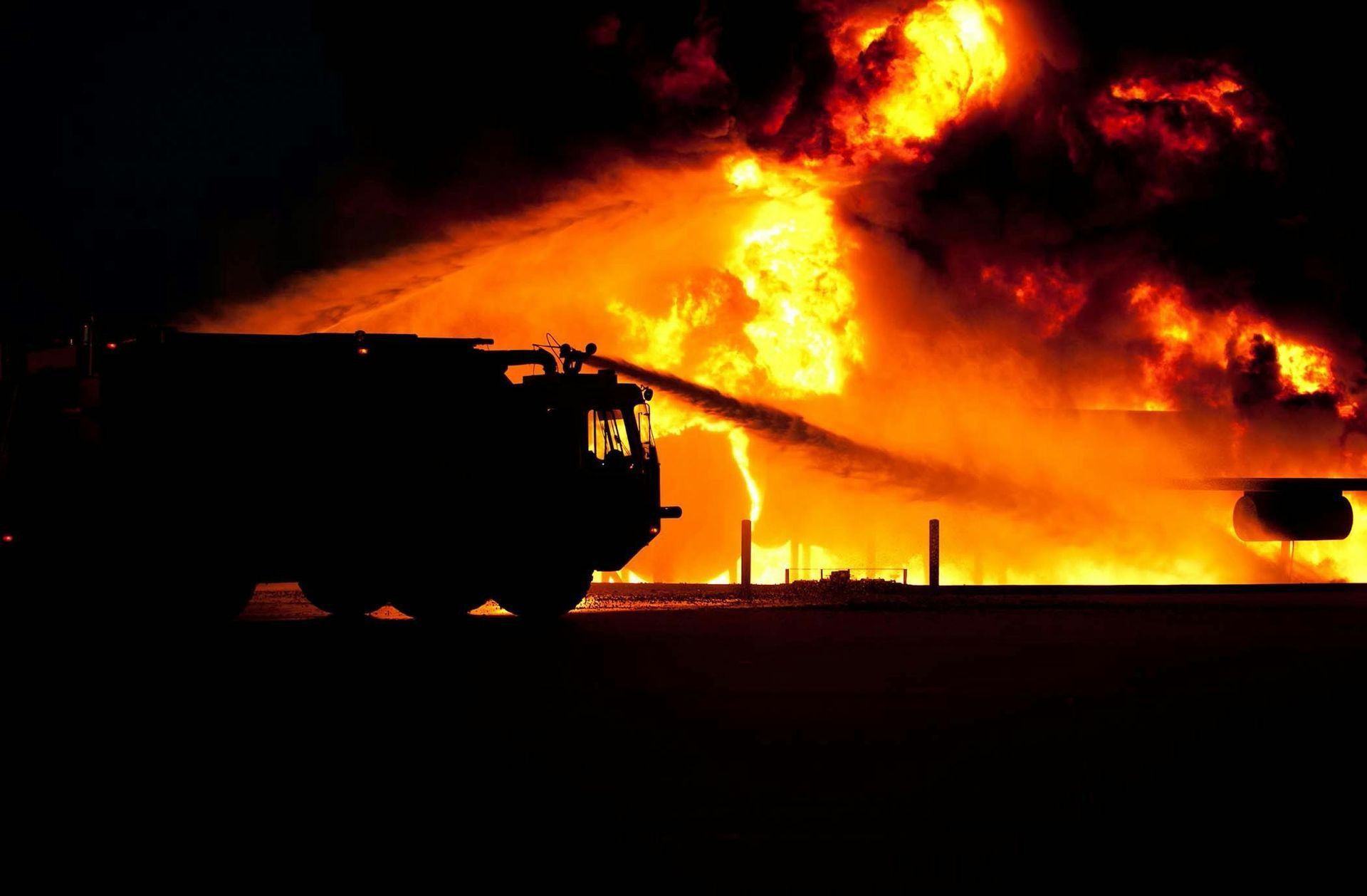 Fire truck spraying water on a large, intense fire at night. Orange flames dominate.