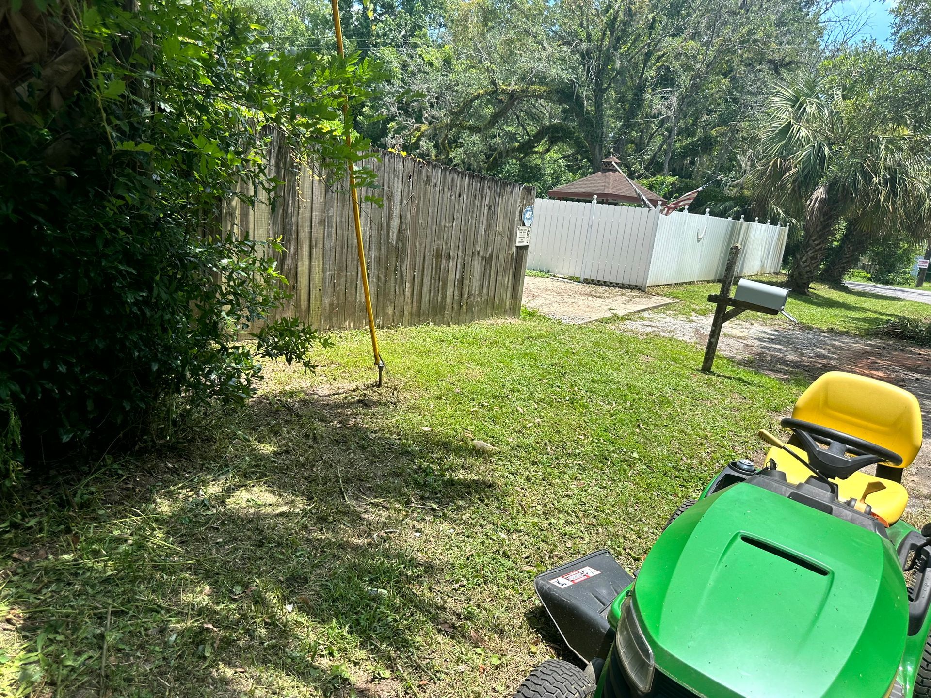 Green riding lawnmower on a grassy yard, near a fence and mailbox, with a white fence in the distance.