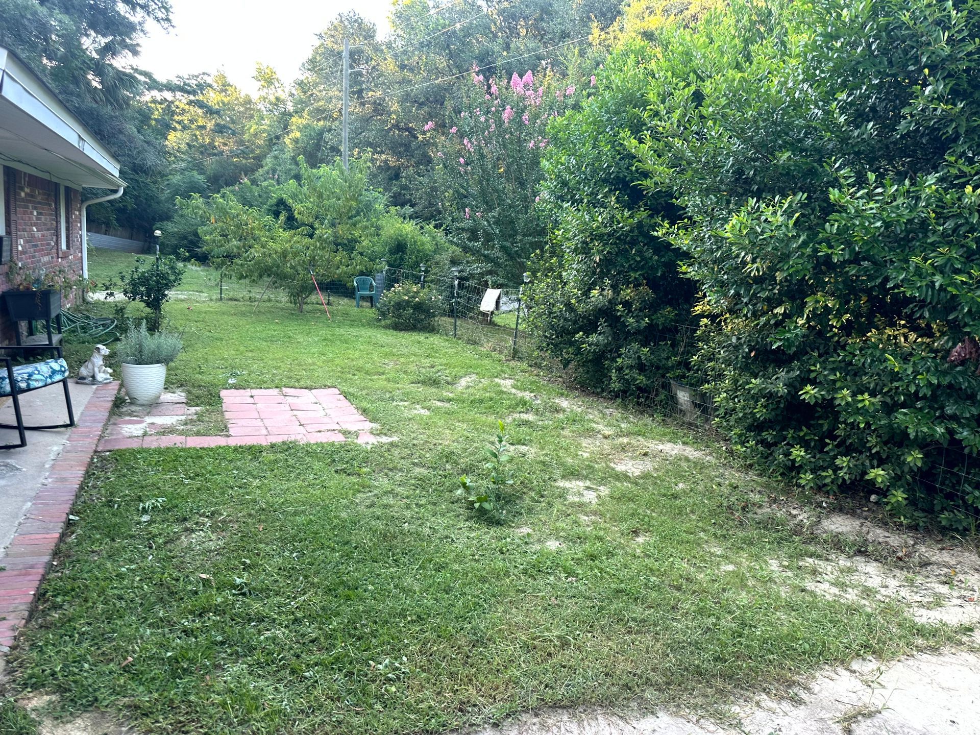 A backyard with patchy green grass, trees, and a brick patio next to a house.
