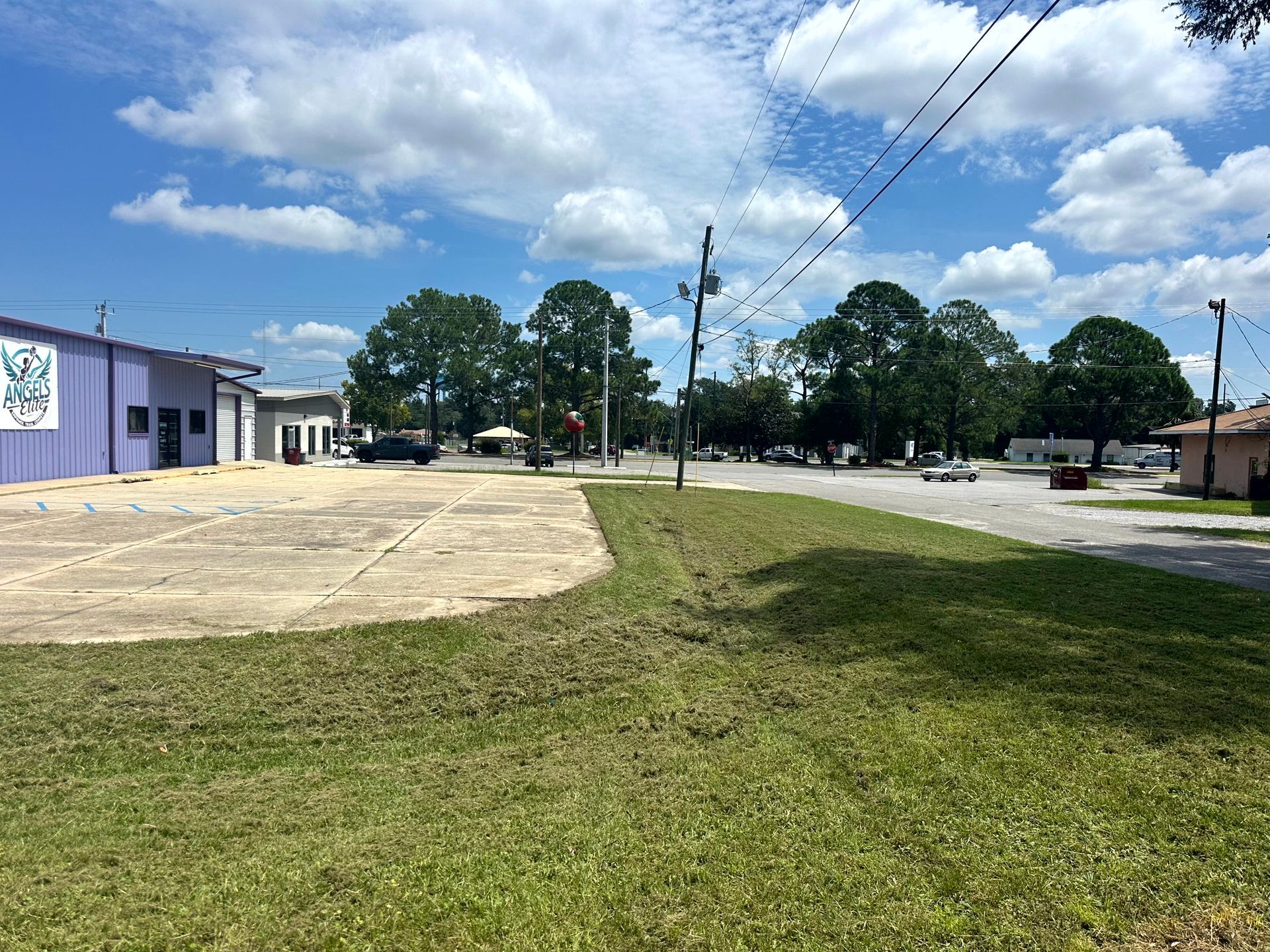 Grassy area leading to a street lined with trees and buildings, under a blue sky with fluffy clouds.