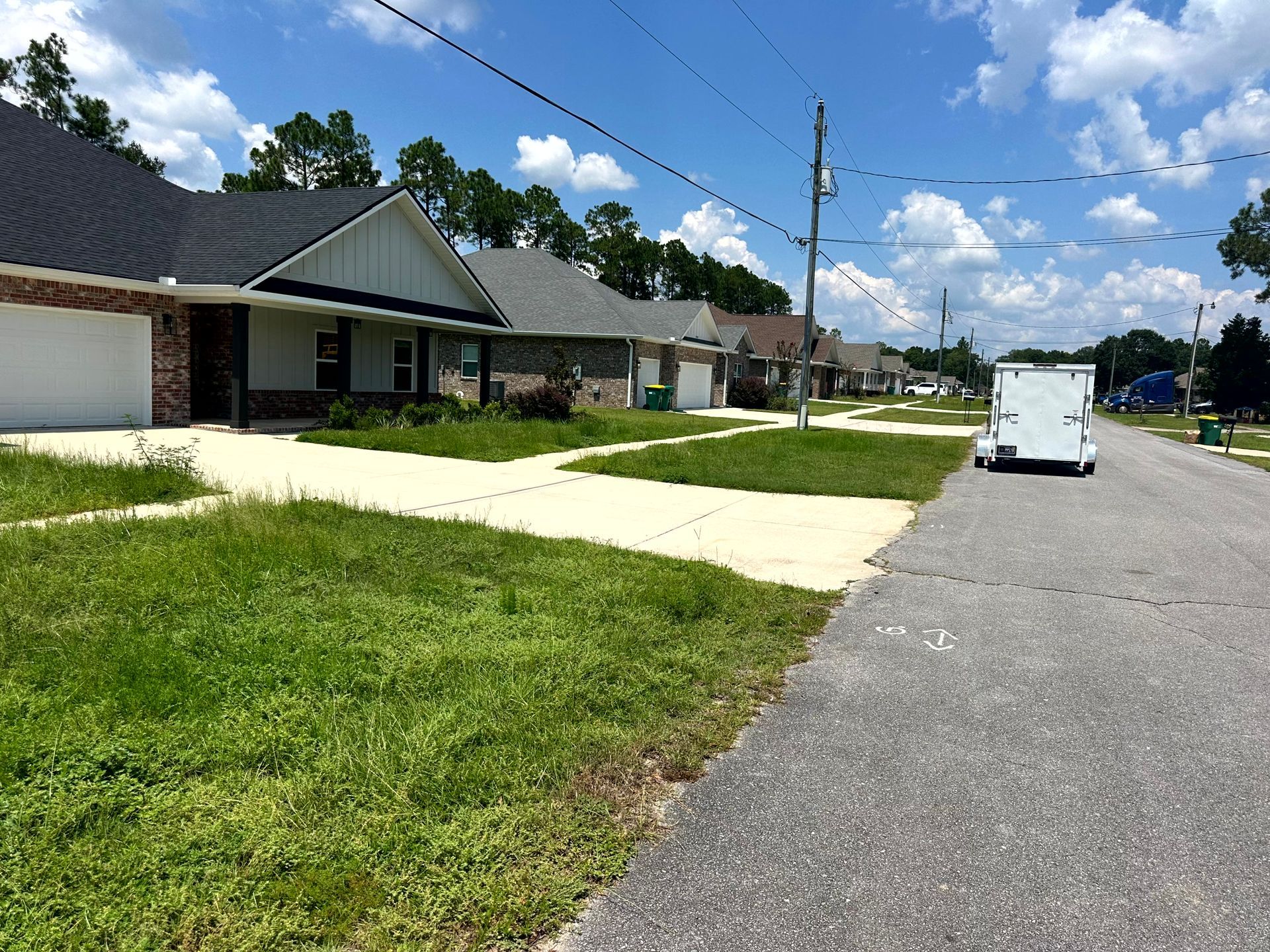 Residential street with houses, overgrown lawns, utility poles, and a trailer on a sunny day.
