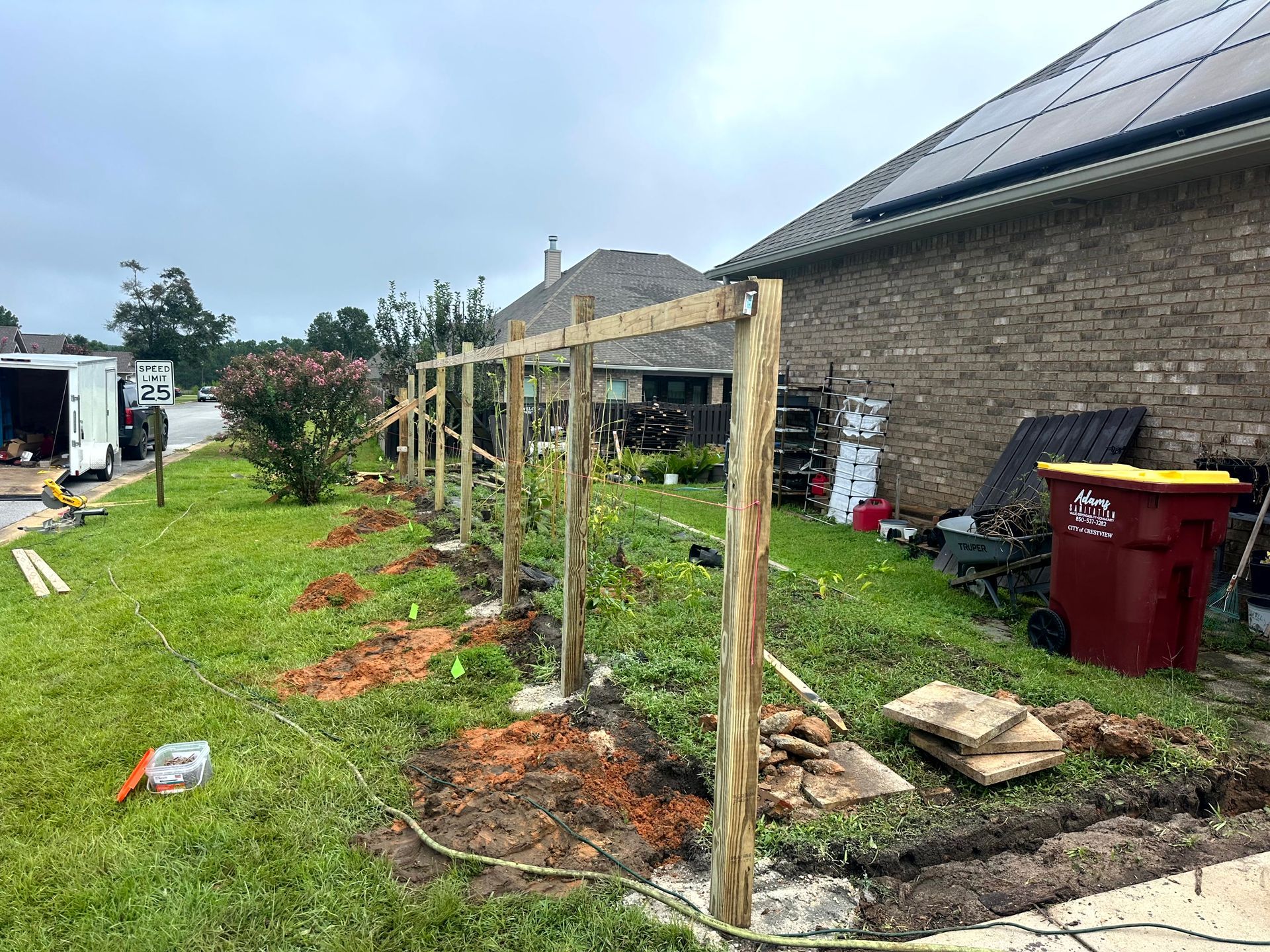 A partially built wooden fence in a yard next to a brick house.  Green grass and brown soil are in the foreground.