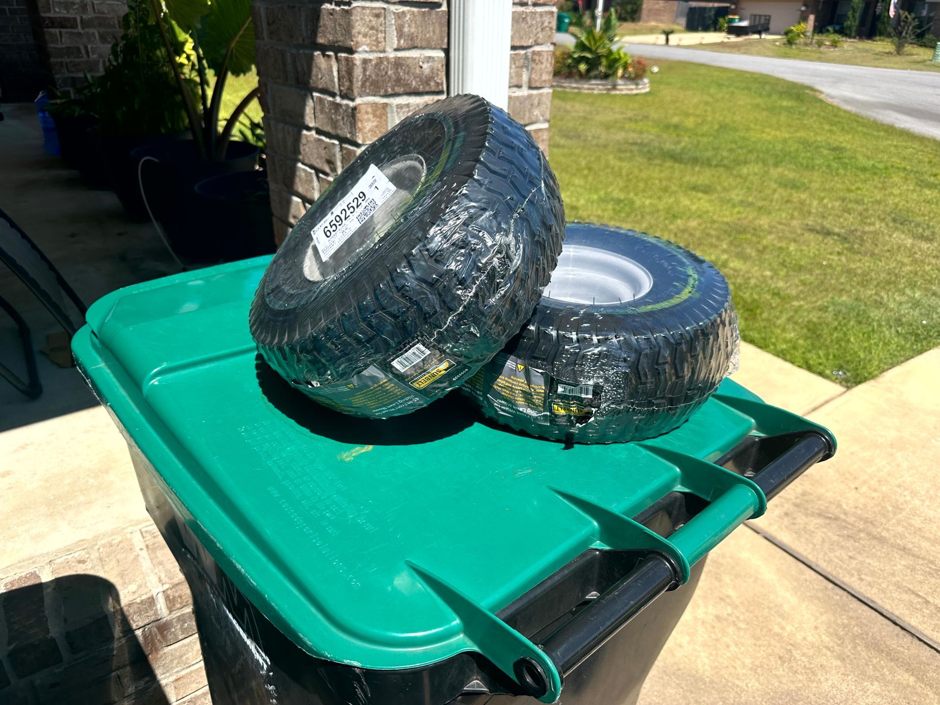 Two new tires wrapped in plastic on top of a green trash bin outdoors.