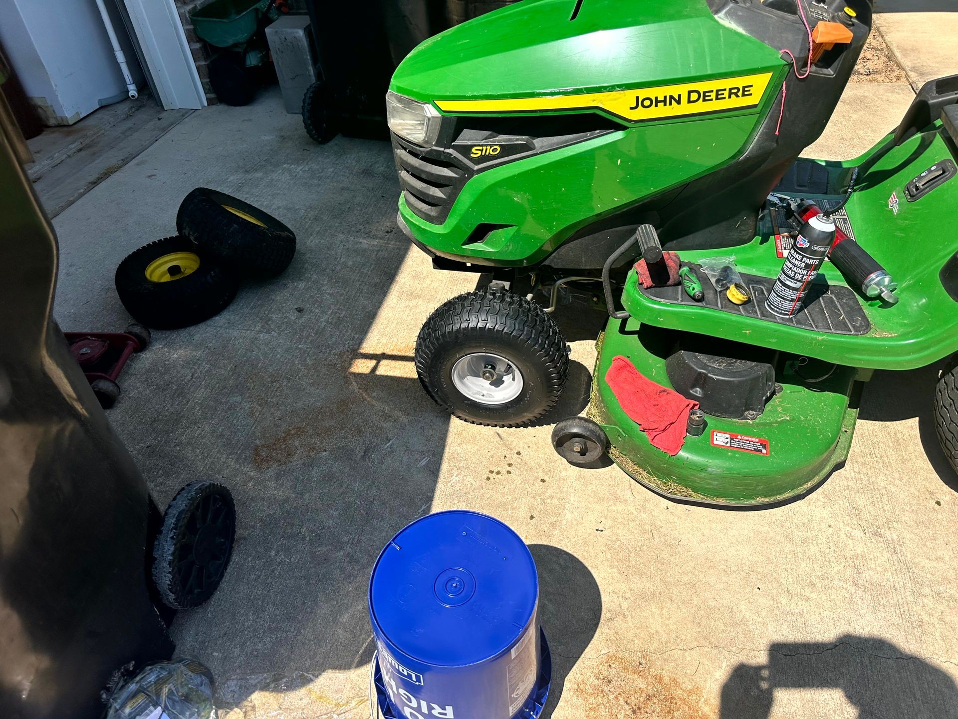 John Deere riding lawn mower with detached tires and blue bucket on a concrete surface.