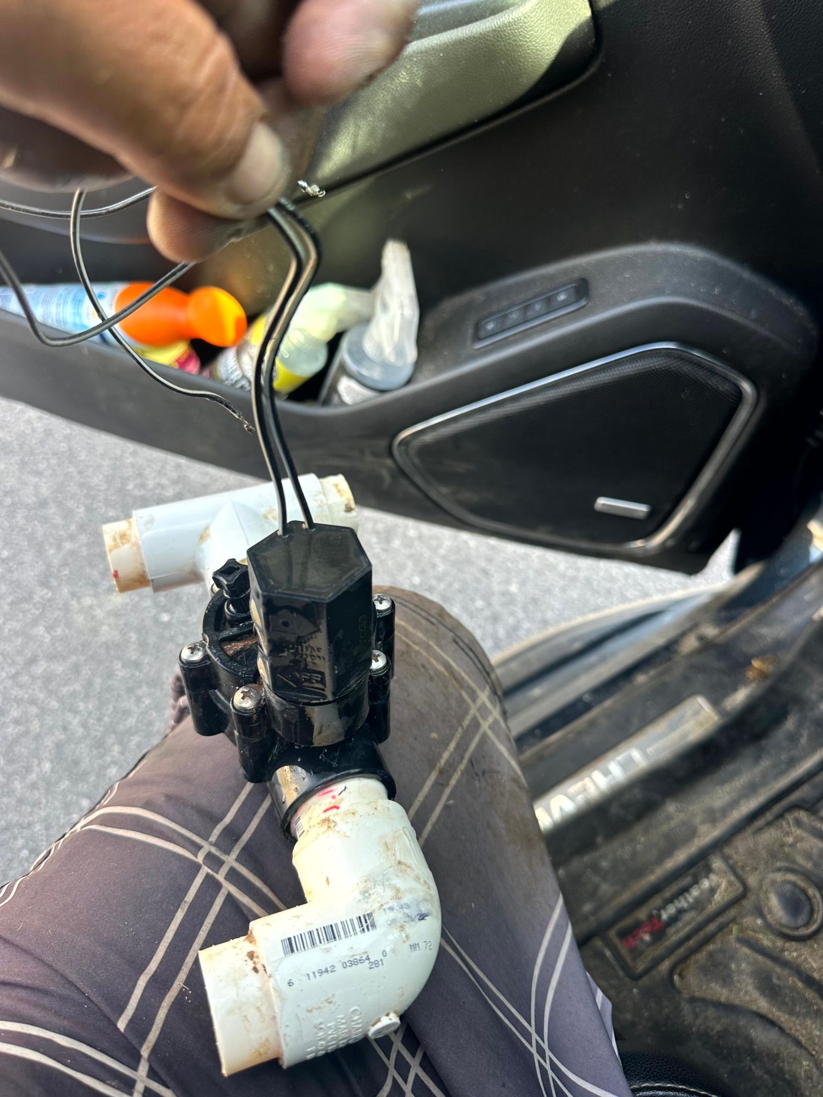 A person holding a black valve with white PVC pipes. Wires attached; setting appears to be inside a car.