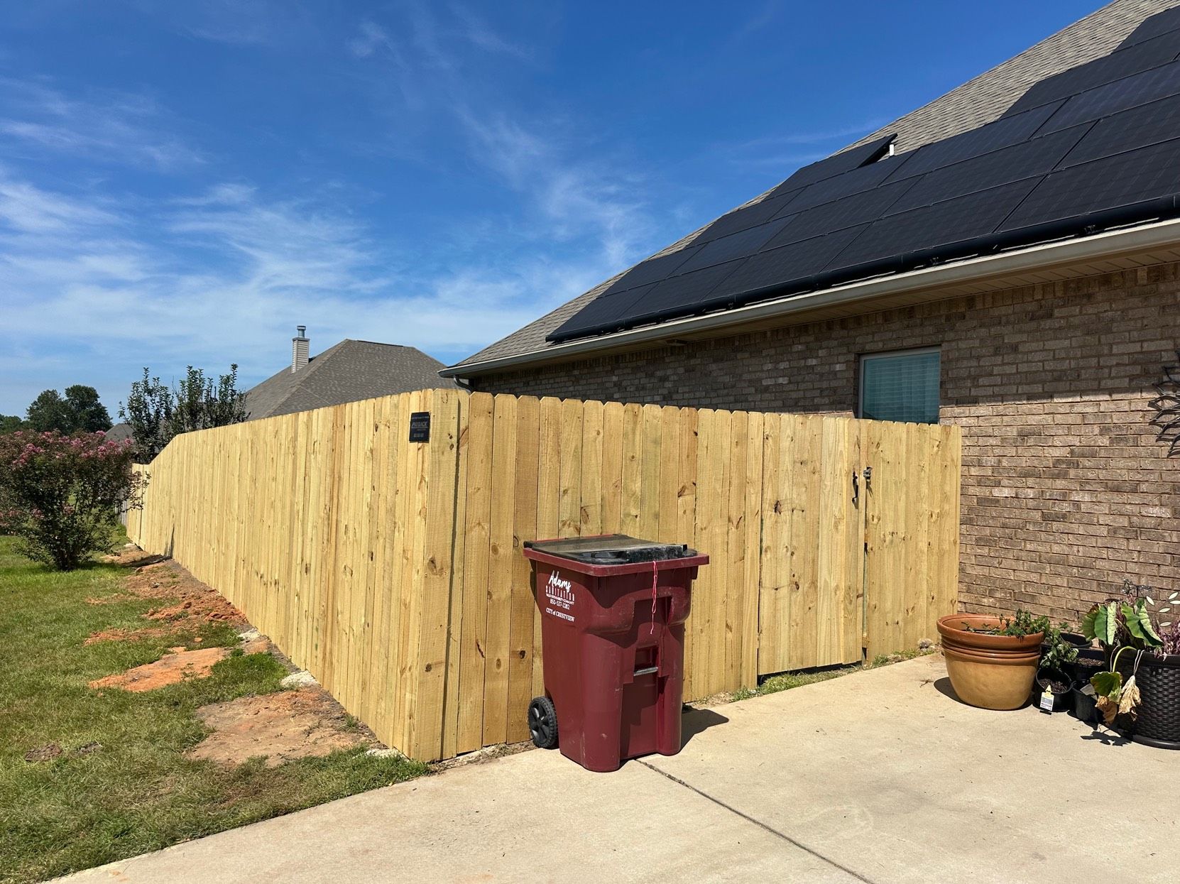 A wooden fence under construction next to a brick house with solar panels on a sunny day.