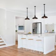 A kitchen with white cabinets , stools , a refrigerator and a staircase.