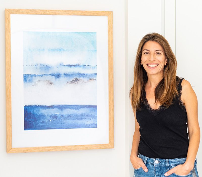 A woman is standing next to a framed painting on a wall.