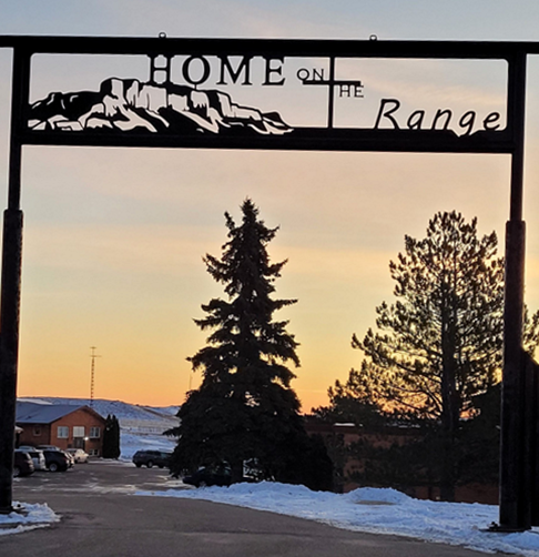 A metal gate sign reading "HOME ON THE RANGE" frames a snowy landscape at sunrise, featuring two large evergreen trees.