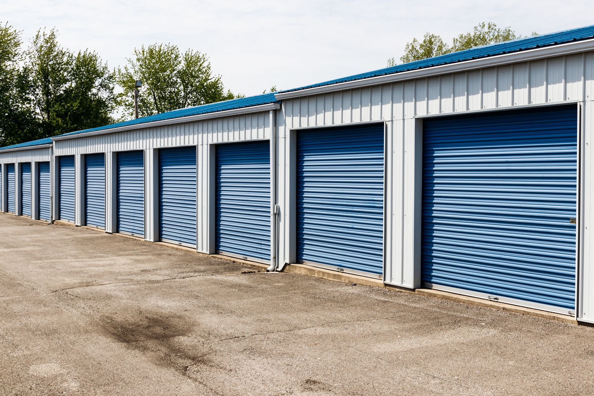 A row of self-storage units with blue roll-up doors against a light building exterior and green trees under a clear sky.