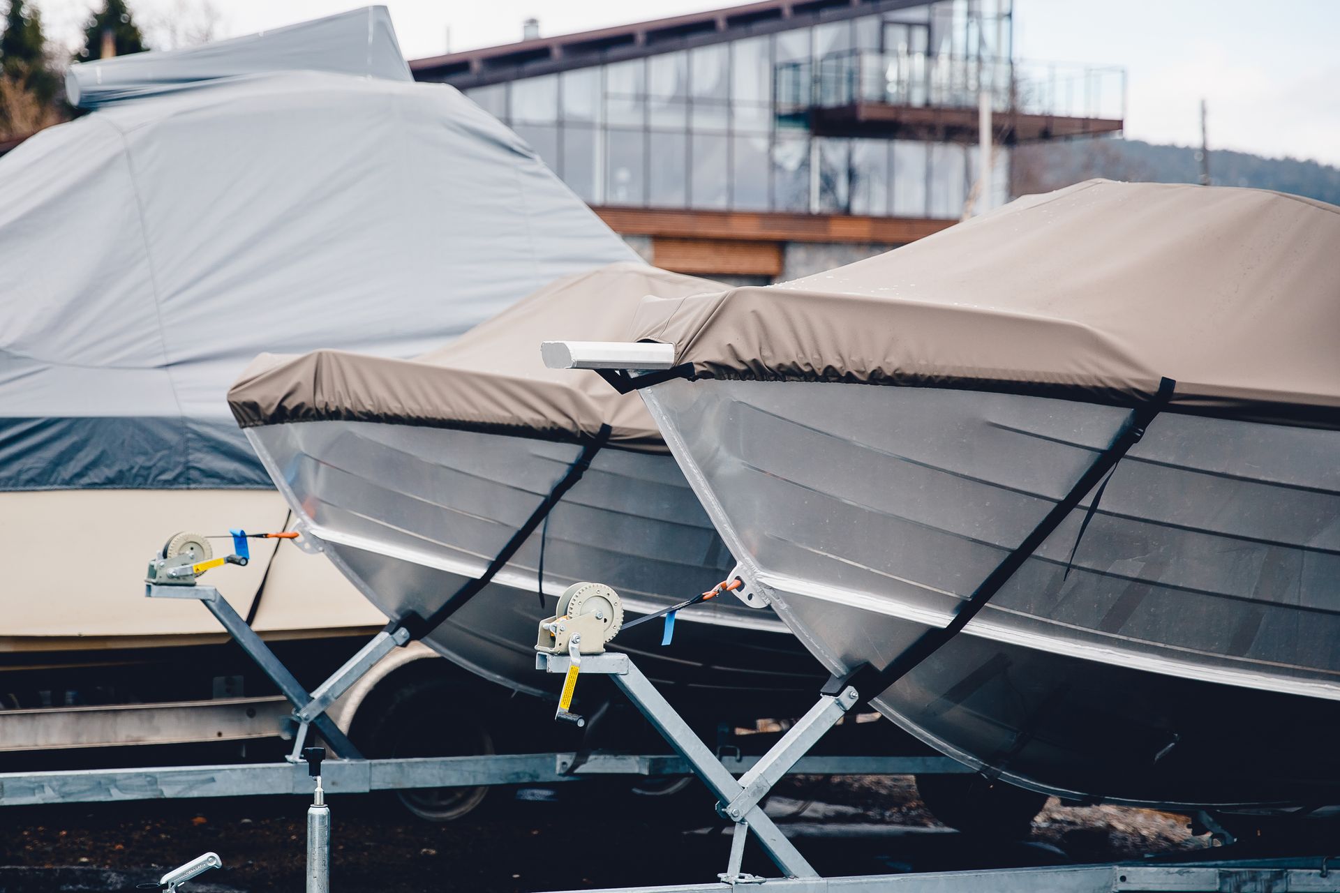 Two boats covered with tan and gray tarps on boat trailers parked in front of a building.