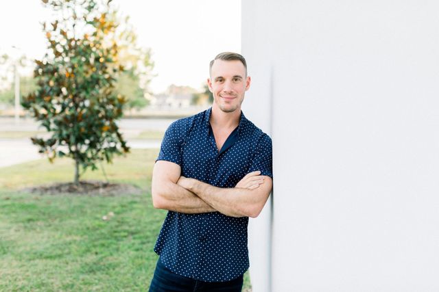 A man in a blue shirt is leaning against a white wall with his arms crossed.