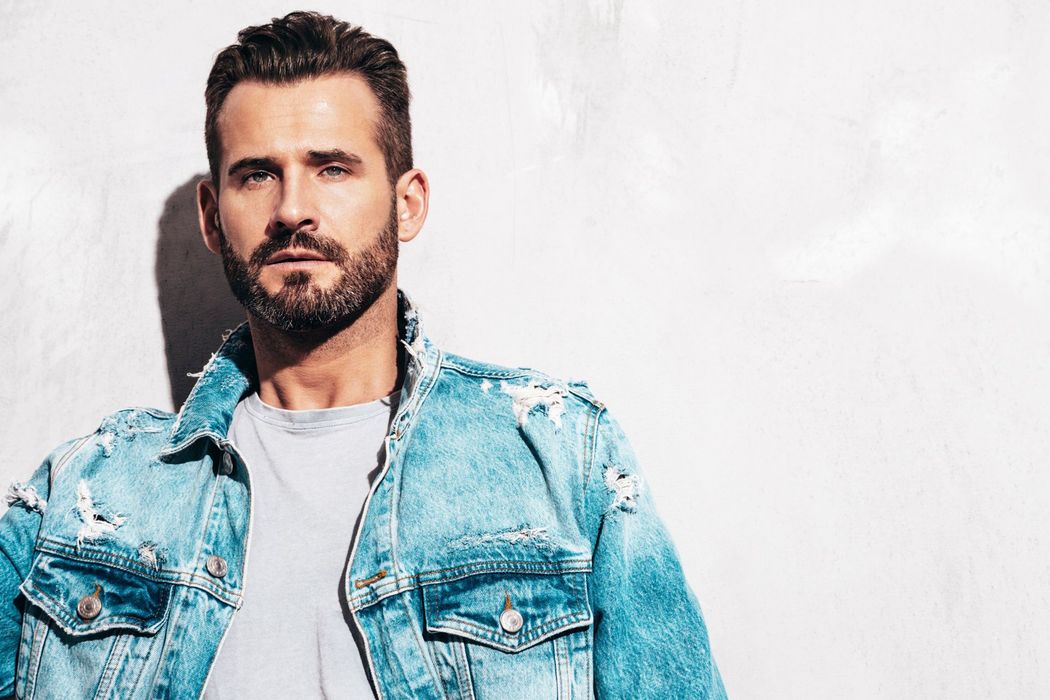 A man with a beard is wearing a denim jacket and standing in front of a white wall.