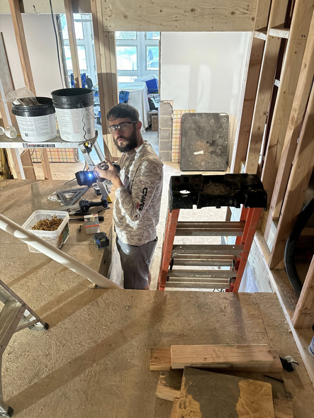 Man in a construction zone, holding tools, looks towards the camera. Wooden framing surrounds the opening.