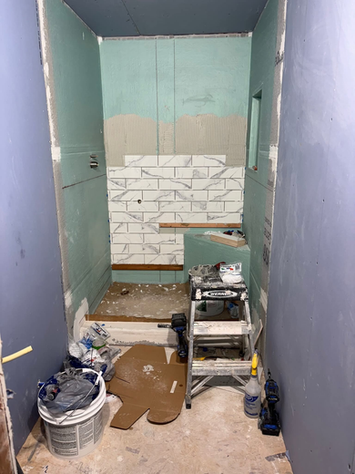 Bathroom under renovation. Walls are green and blue. White and marble tile being installed. Ladder and tools present.