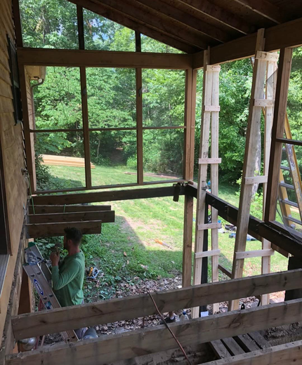 Construction in progress on a screened-in porch with a person working. Wooden beams, scaffolding, and green foliage are visible.