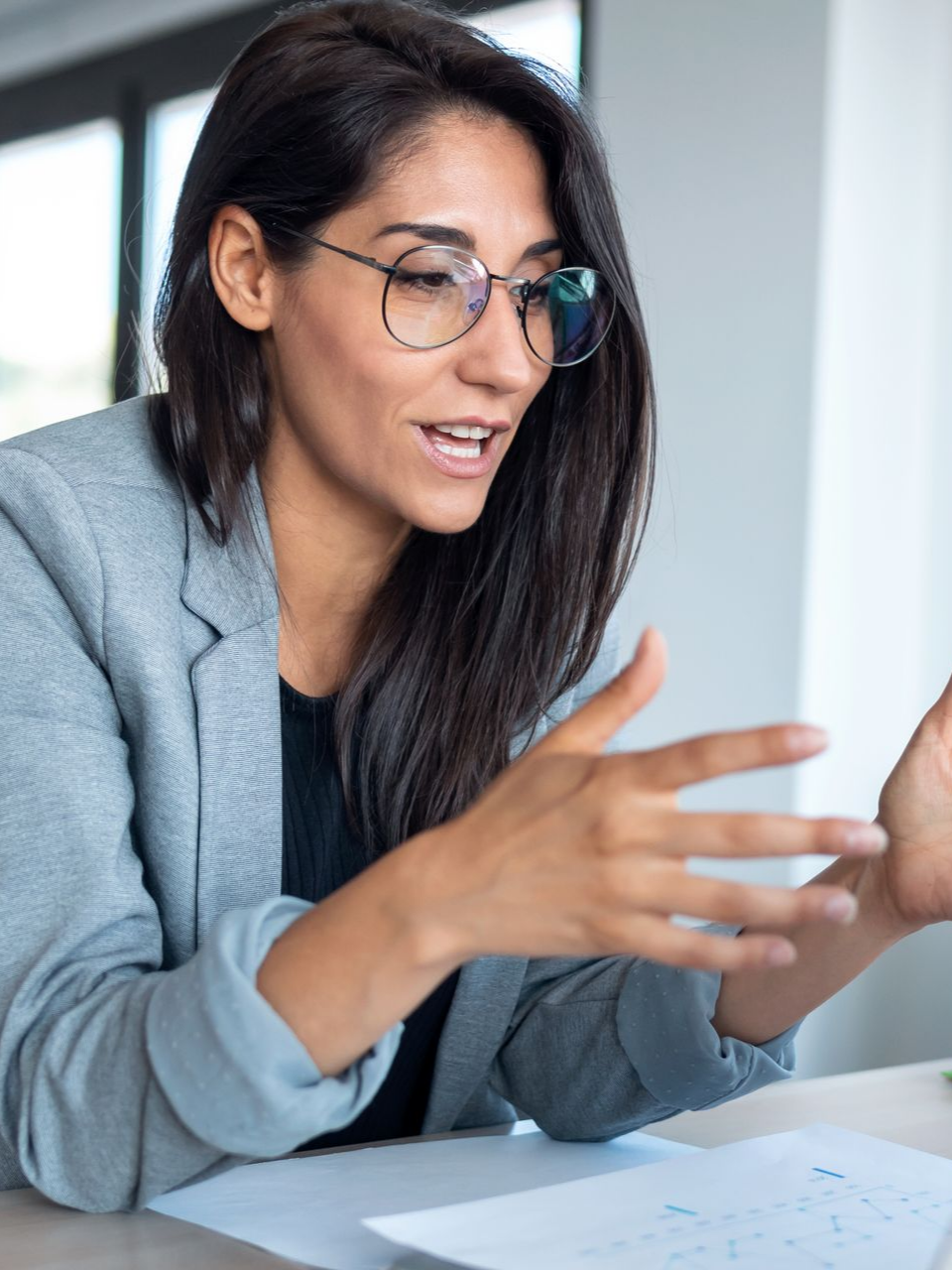Woman in glasses, gray blazer, gesturing with hands during a discussion.