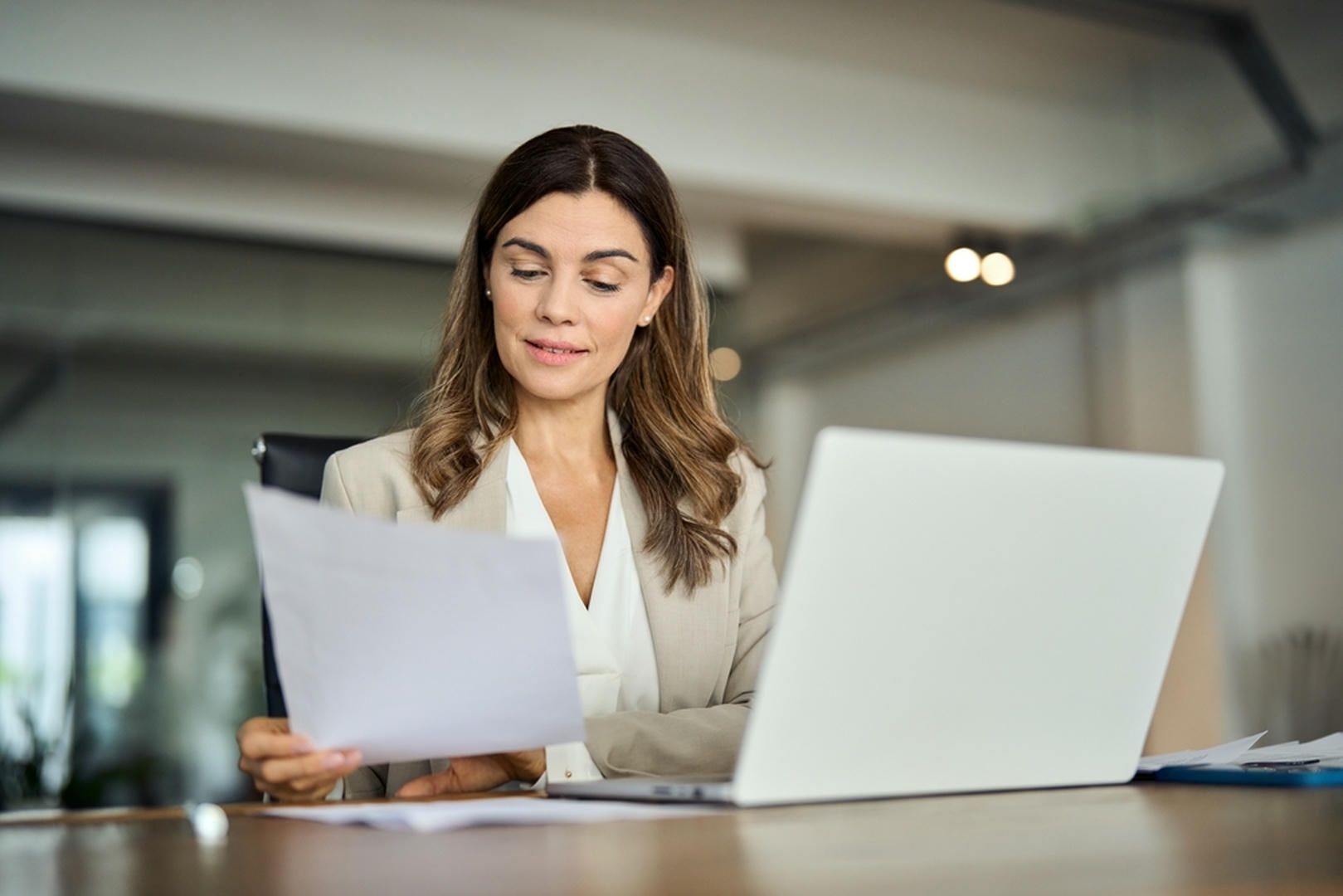 Woman in a blazer seated at a desk, looking at papers while using a laptop.