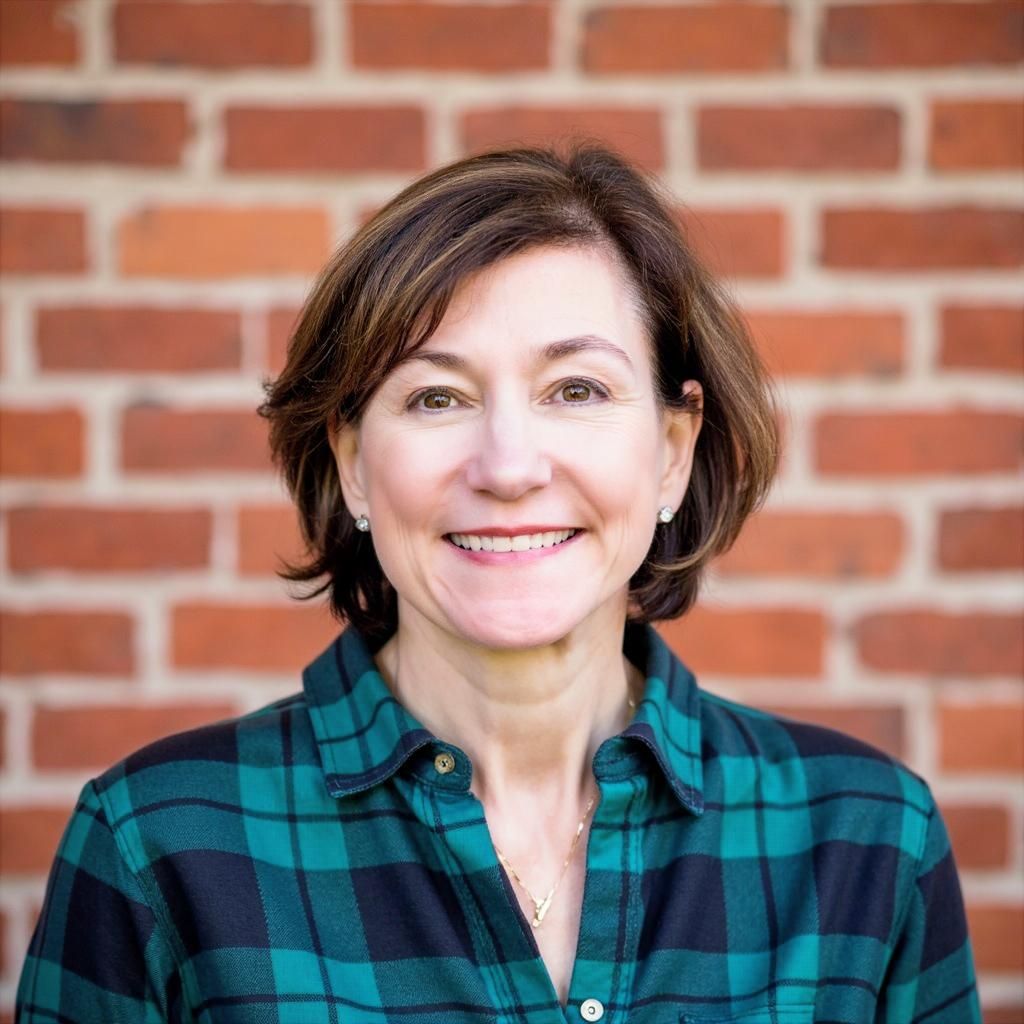 Woman with short brown hair smiles, wearing a green and black plaid shirt in front of a brick wall.