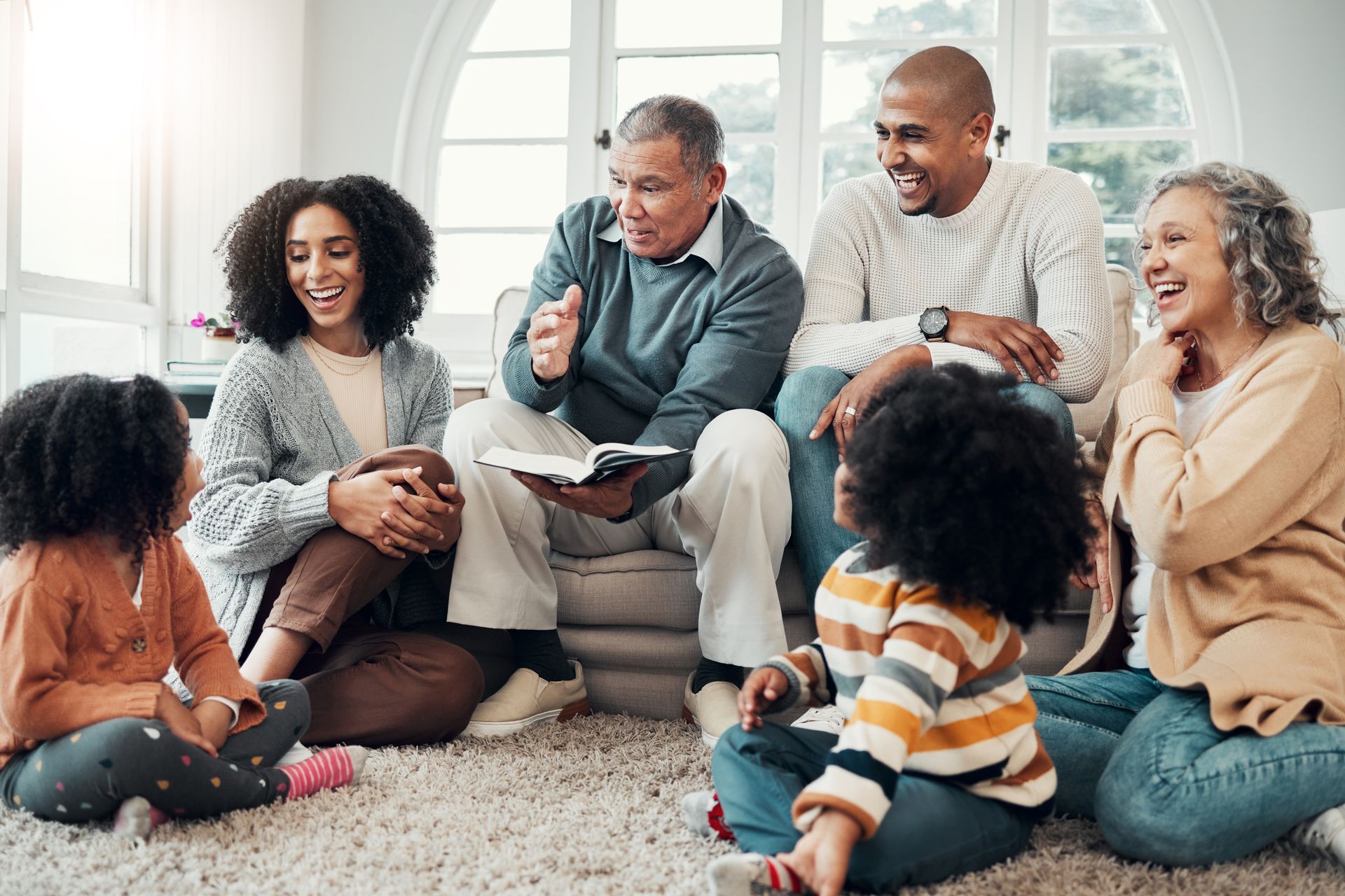 Family sitting on the floor, reading and laughing together in a bright living room.