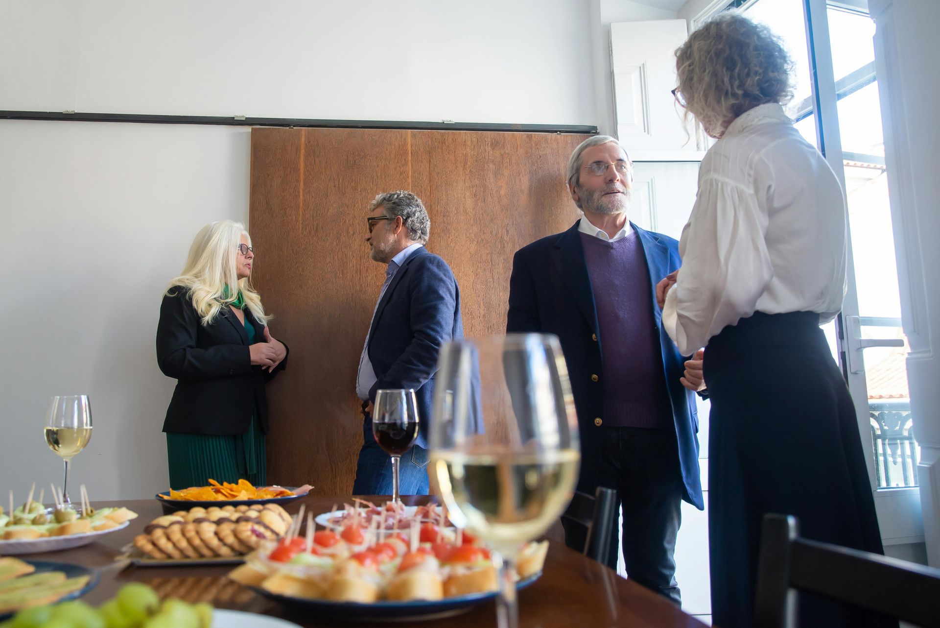 People conversing near a buffet table with appetizers and wine glasses. Light-filled room, some wearing jackets.