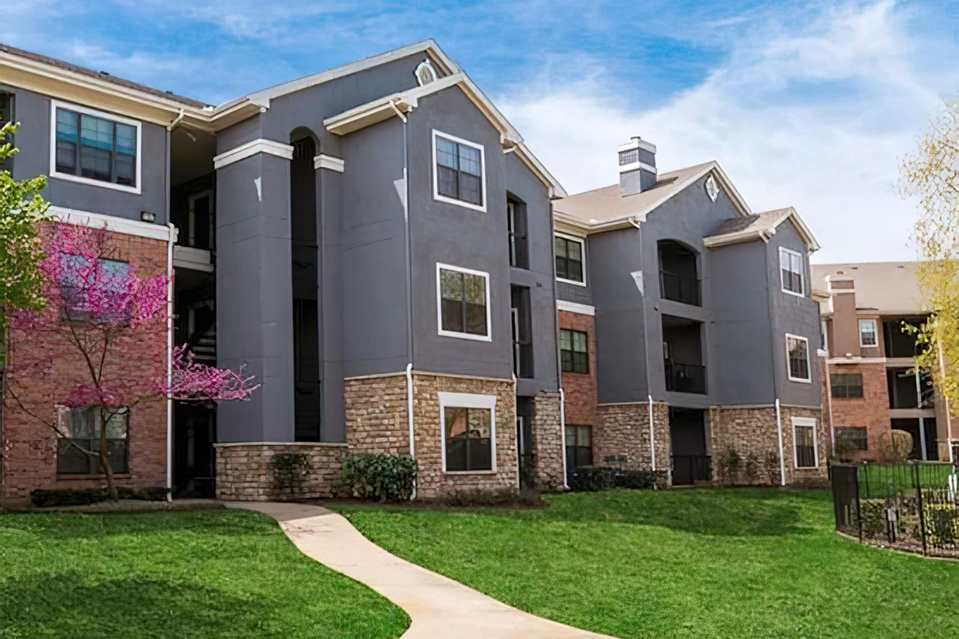 Apartment complex with gray and tan exteriors, surrounded by green grass and trees under a blue sky.