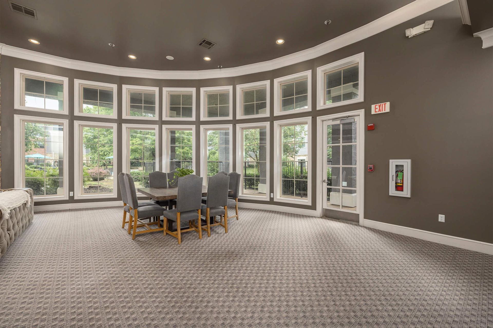 A dining area with a round table, chairs, and many windows, with a carpeted floor and gray walls.