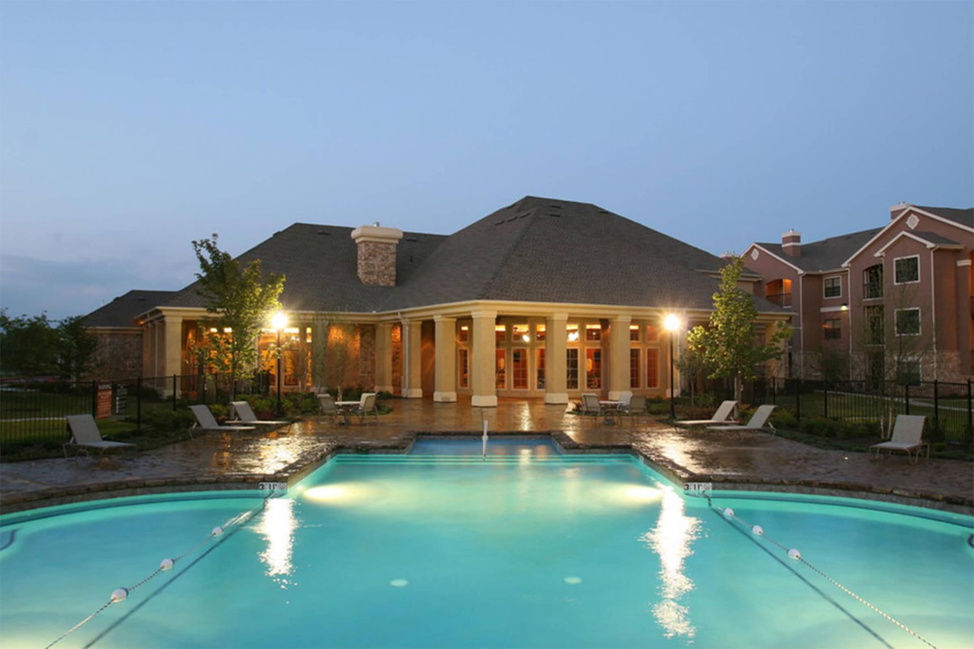 Pool at dusk in front of a building with pillars; lounge chairs lined up around the pool.