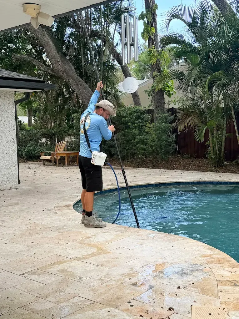 Person cleaning a pool with a long pole on a patio beside palm trees