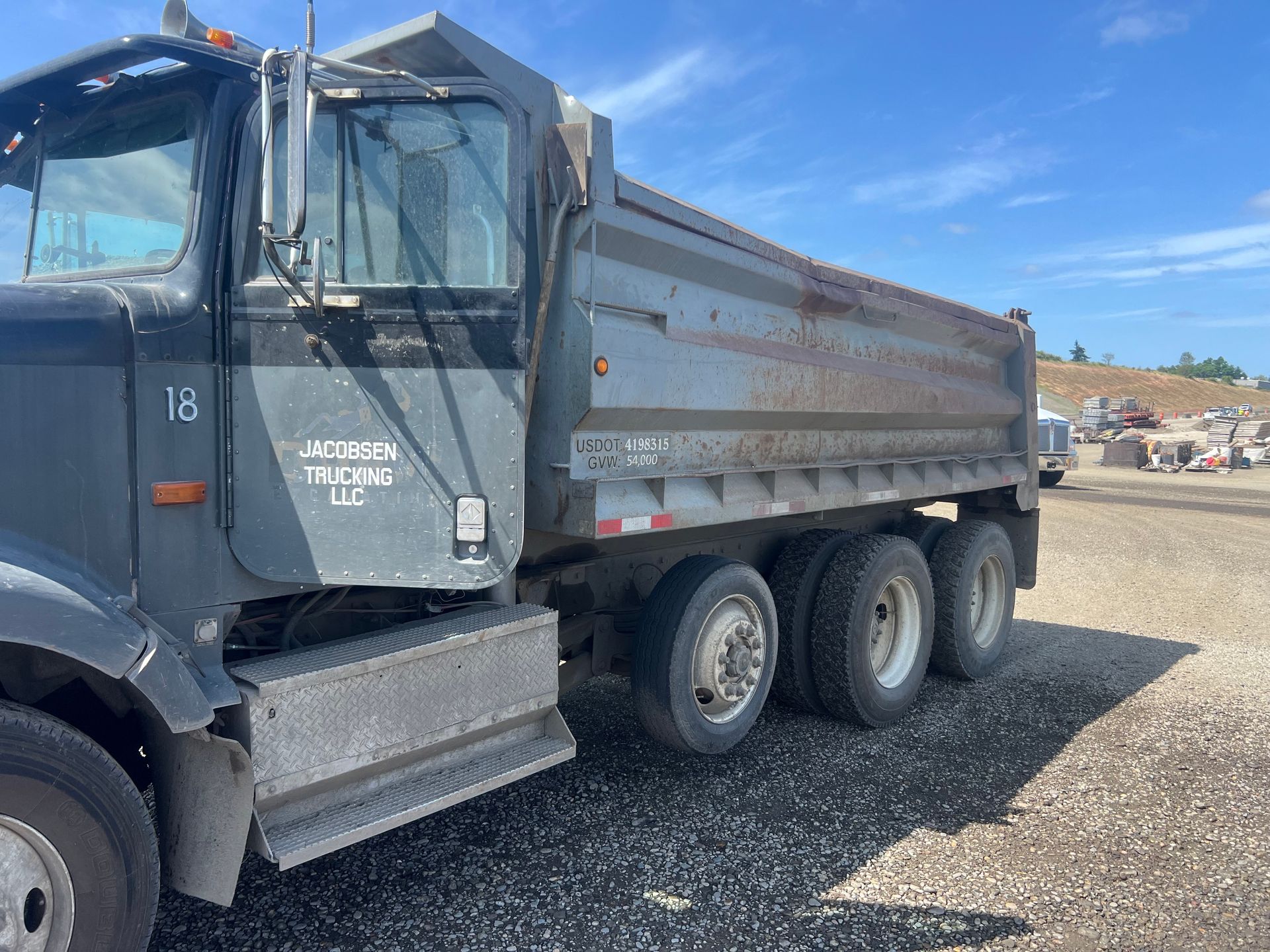 A grey dump truck filled with gravel parked on cobblestones, with a red car and construction vehicle visible.