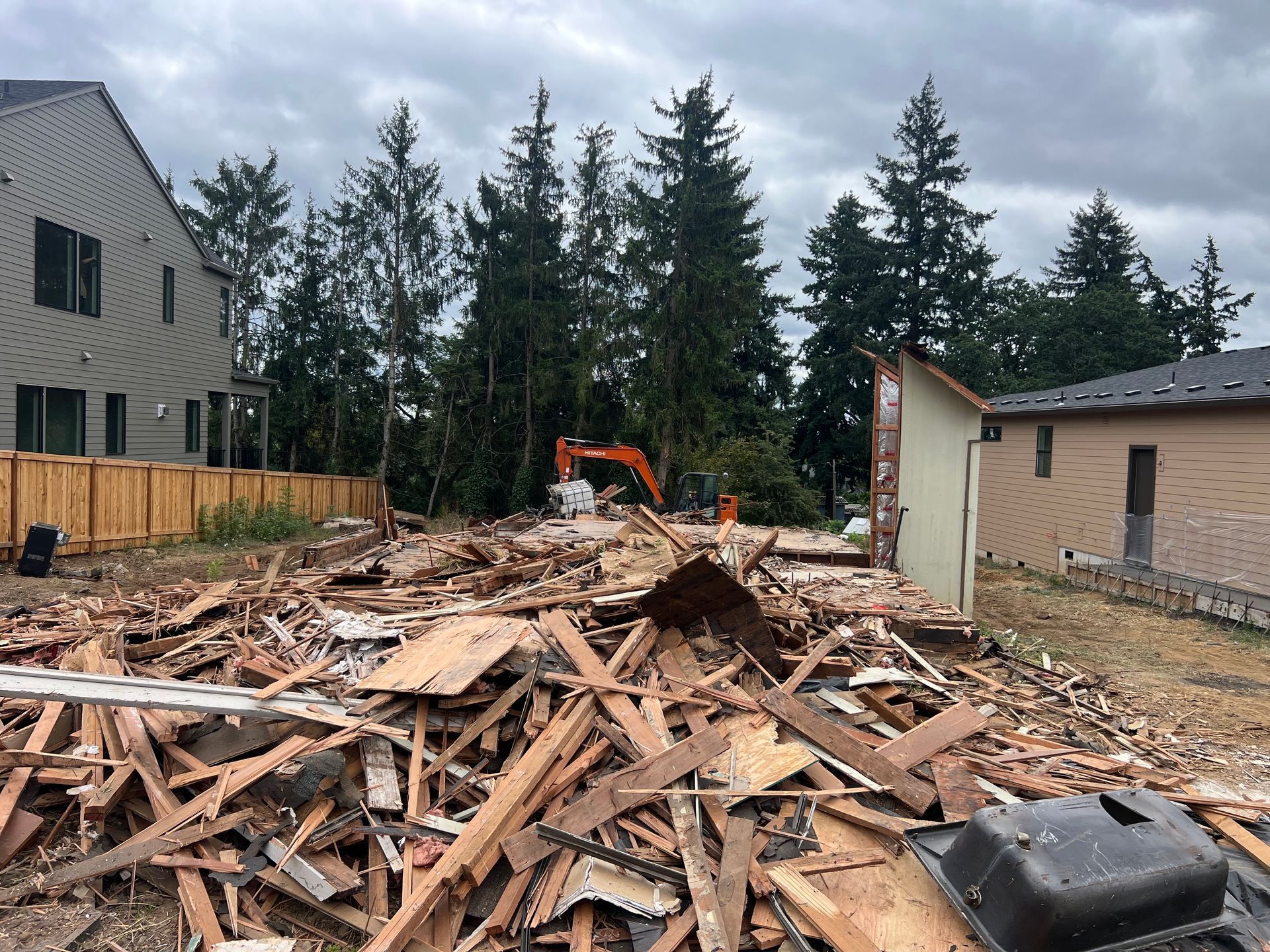 An excavator demolishes a brick building, kicking up dust. The machine is orange, and debris litters the site.