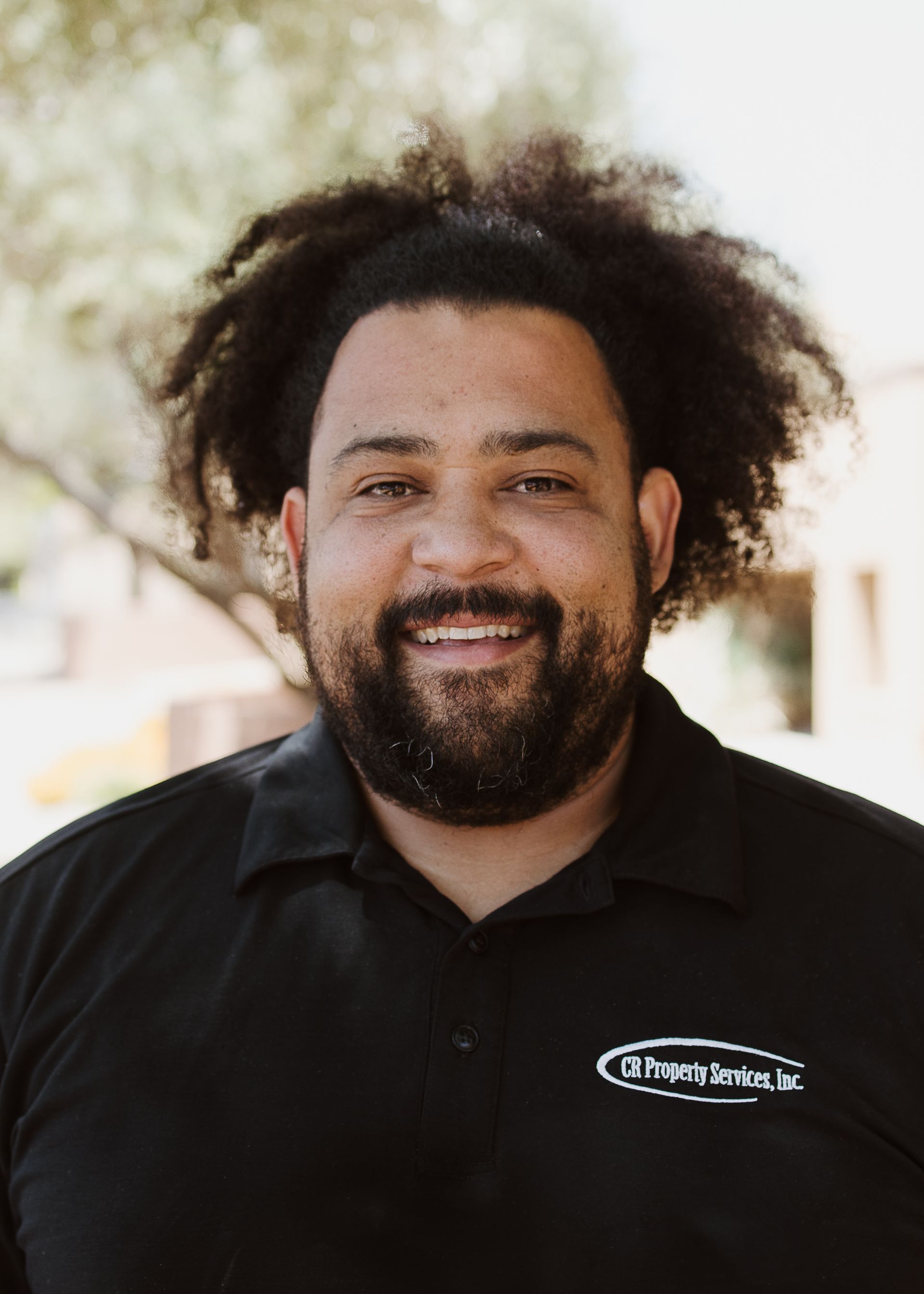 A man with curly hair and a beard is wearing a black shirt and smiling.