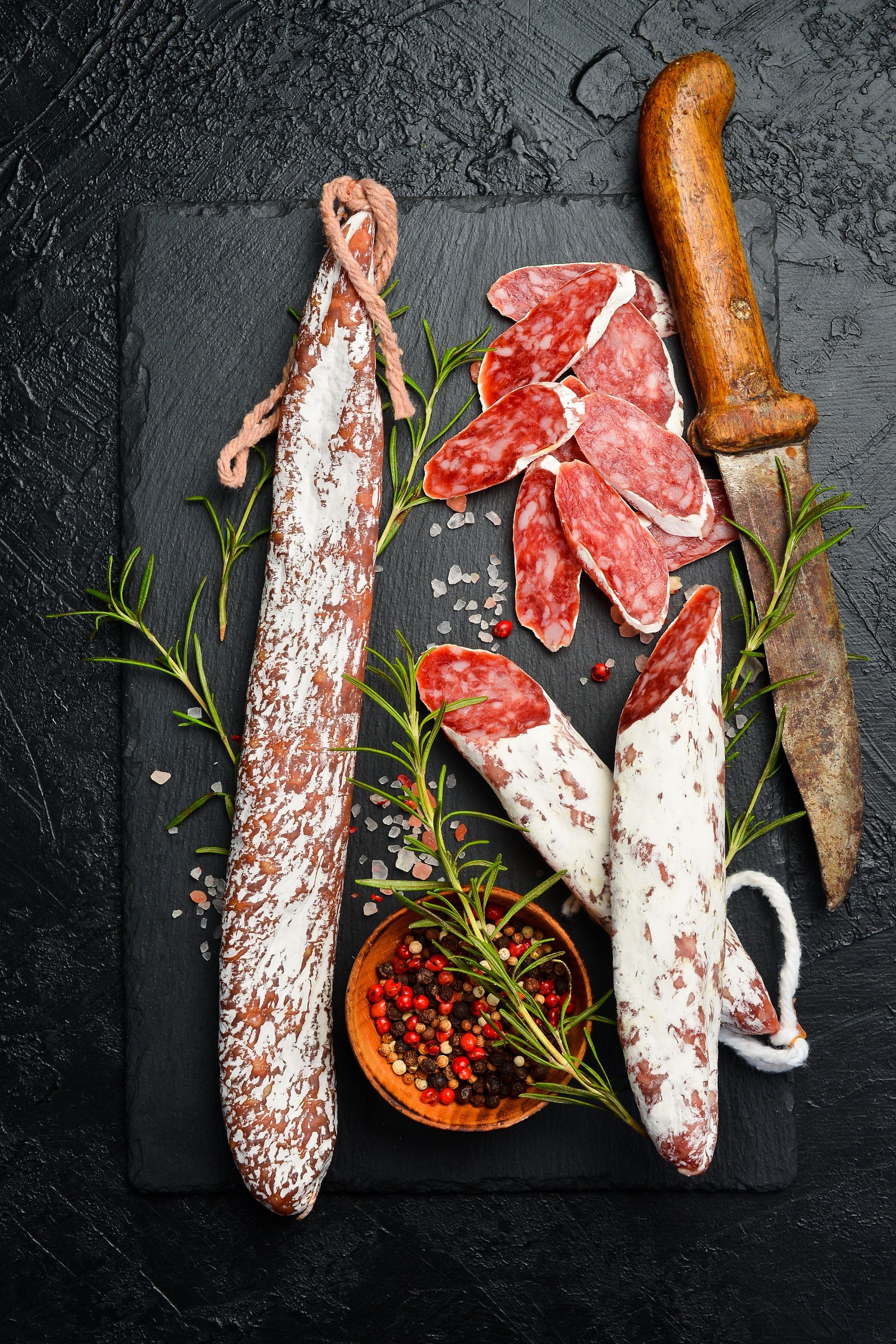 Salami auf schwarzem Schiefer mit einem Messer, Rosmarin und Pfefferkörnern.