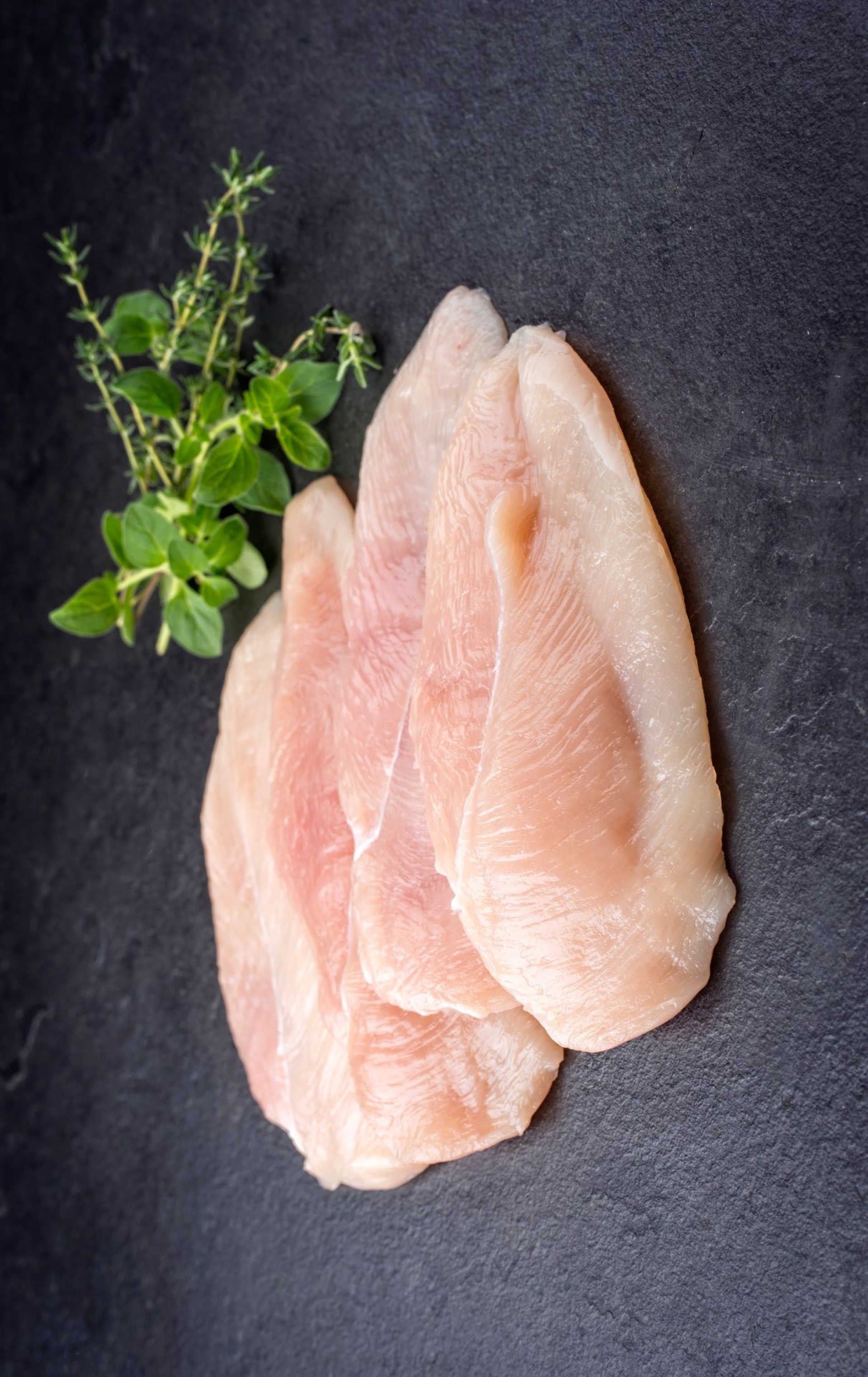 Drei rohe Hähnchenbrüste und ein Zweig grüner Kräuter auf einer dunklen Oberfläche.