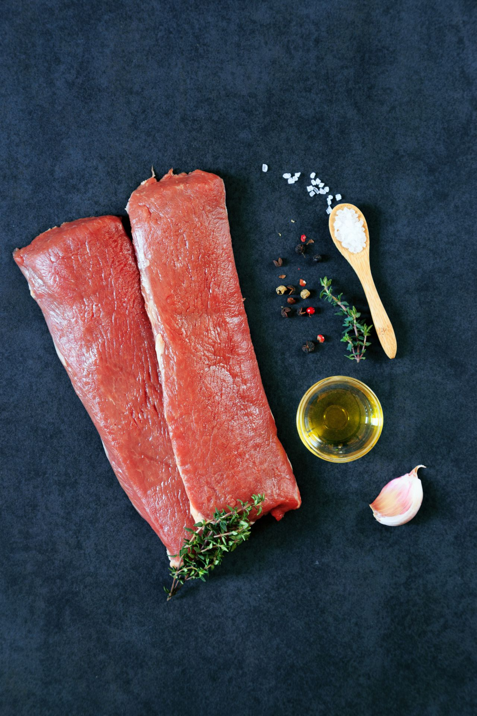 Rohe Lammstücke mit Salz, Pfeffer, Kräutern, Olivenöl und Knoblauch auf einer dunklen Oberfläche.