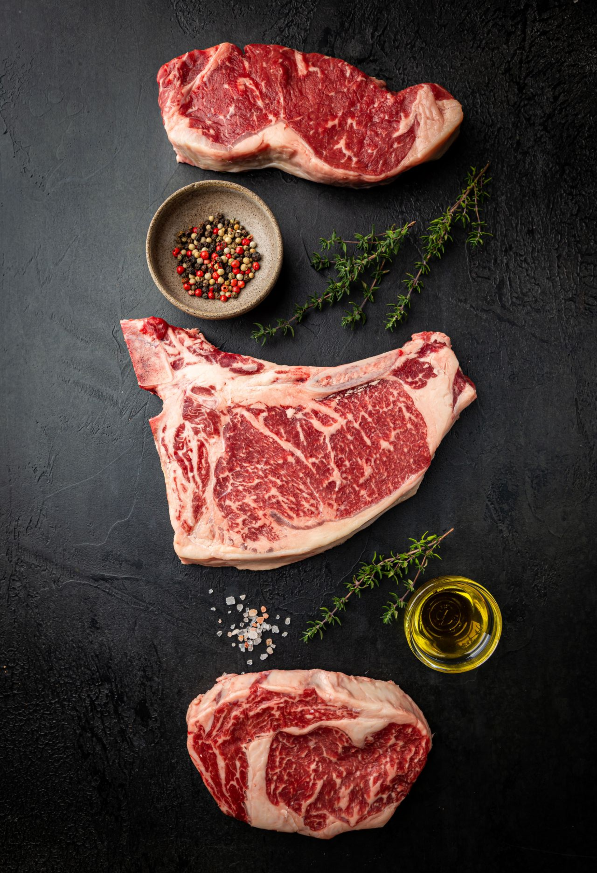 Rohe Steaks, Gewürze, Kräuter und Olivenöl auf einer dunklen, strukturierten Oberfläche.