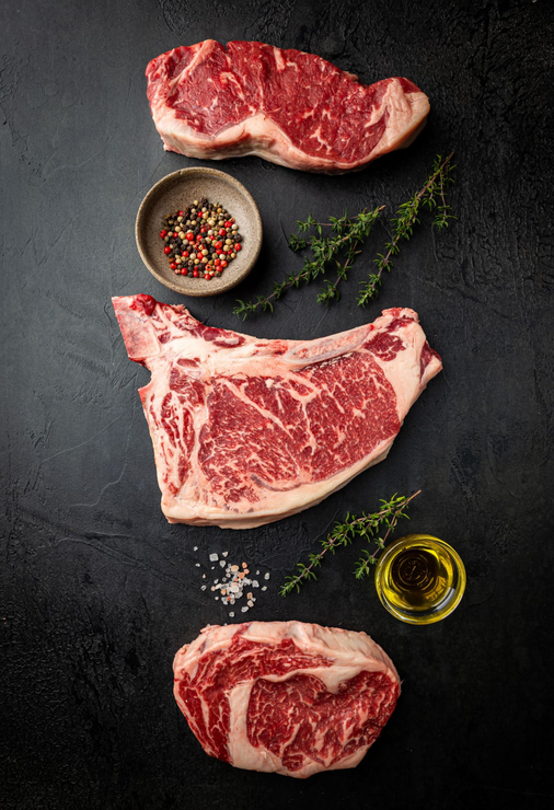 Rohe Steaks, Gewürze, Kräuter und Olivenöl auf einer dunklen, strukturierten Oberfläche.