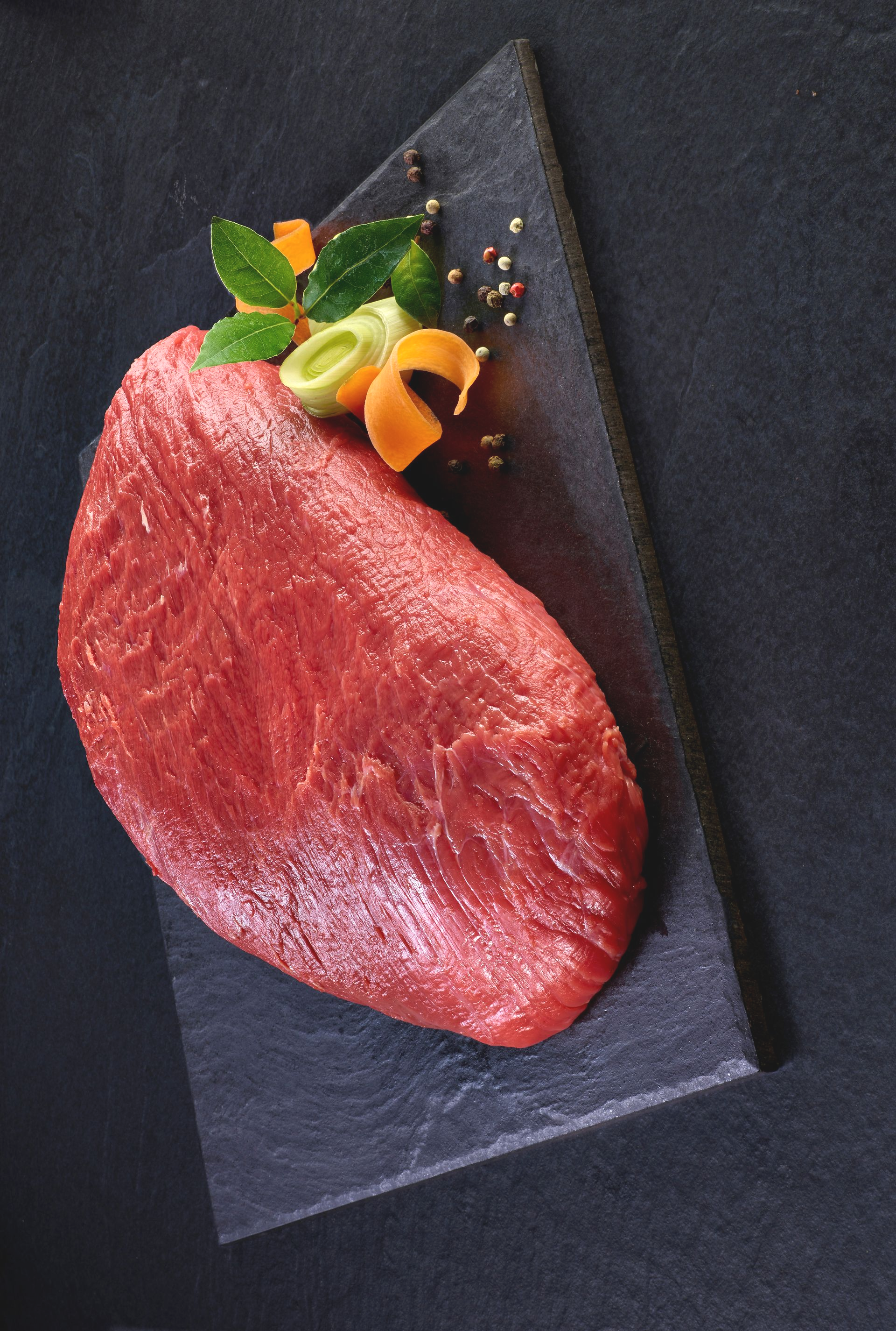 Ein rohes Stück rotes Fleisch auf einem dunklen Schieferbrett, garniert mit Gemüse und Kräutern.
