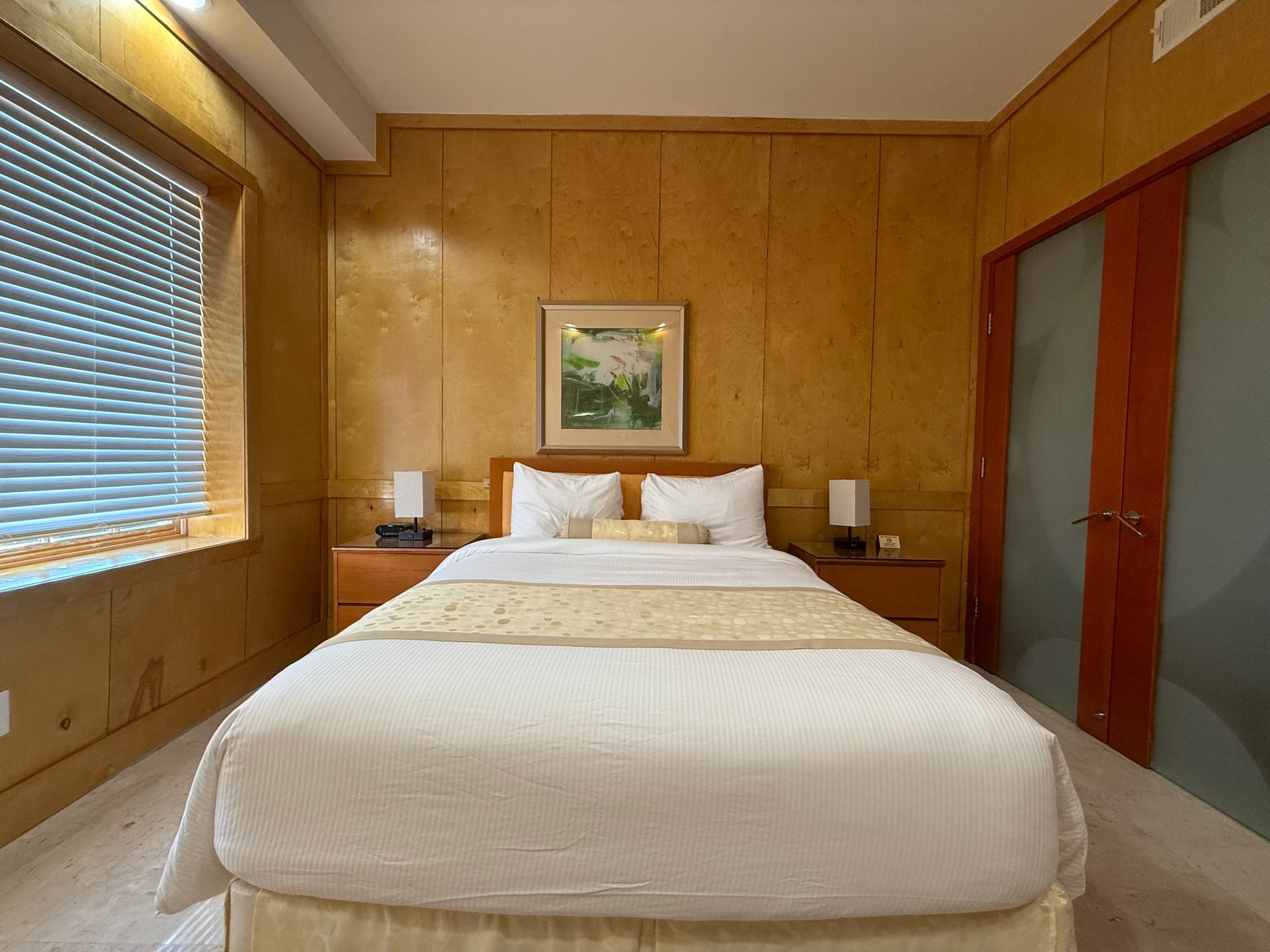 Bedroom with bed, bedside tables, wood-paneled walls, blinds, and a painting.