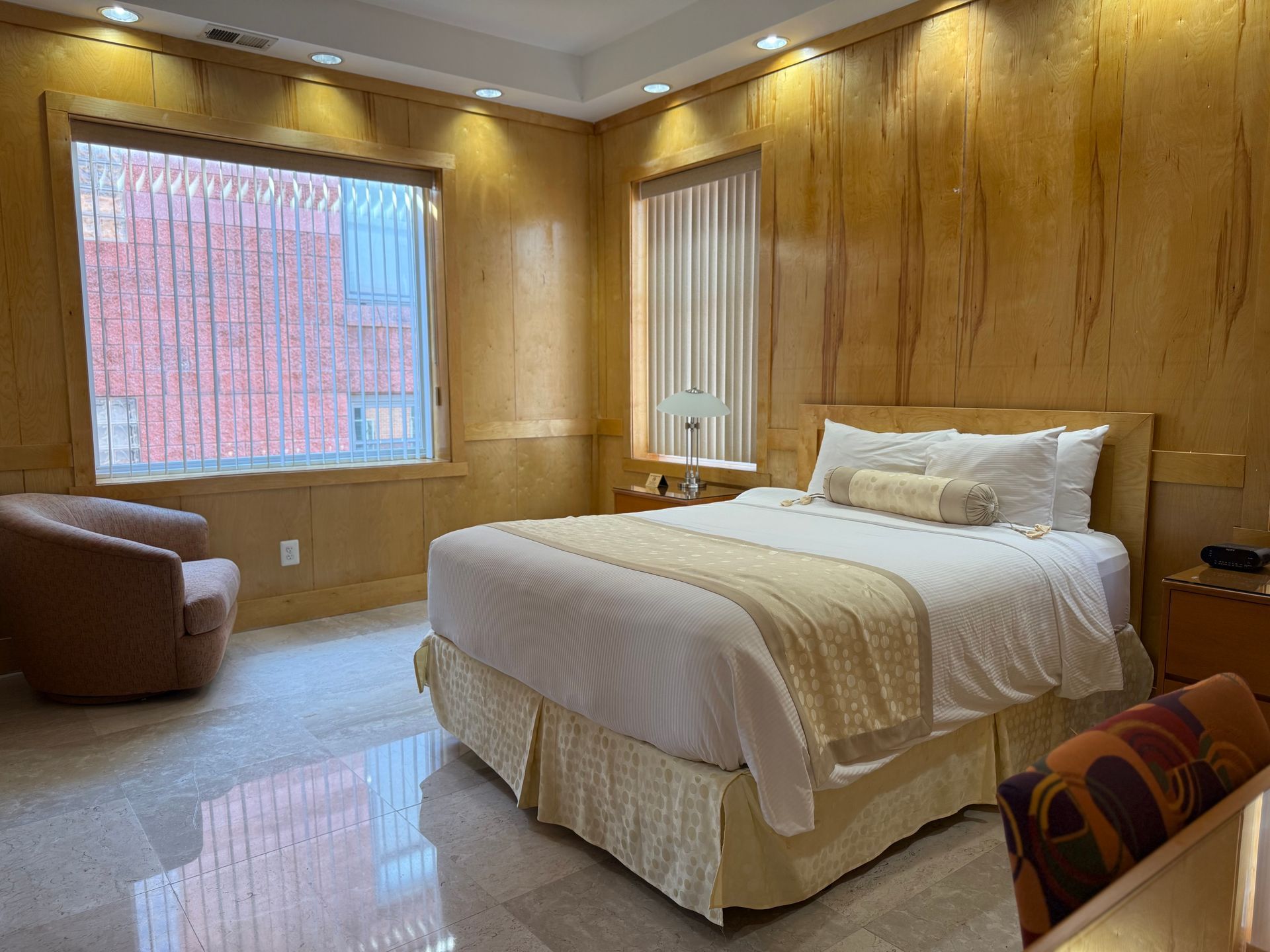 Bedroom with king-size bed, armchair, two windows, and wood-paneled walls, with gold and beige hues.