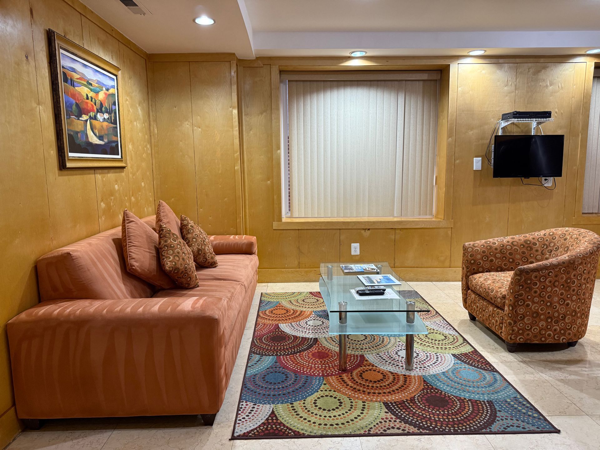 Living room with orange sofa, patterned rug, glass coffee table, and small TV on the wall.