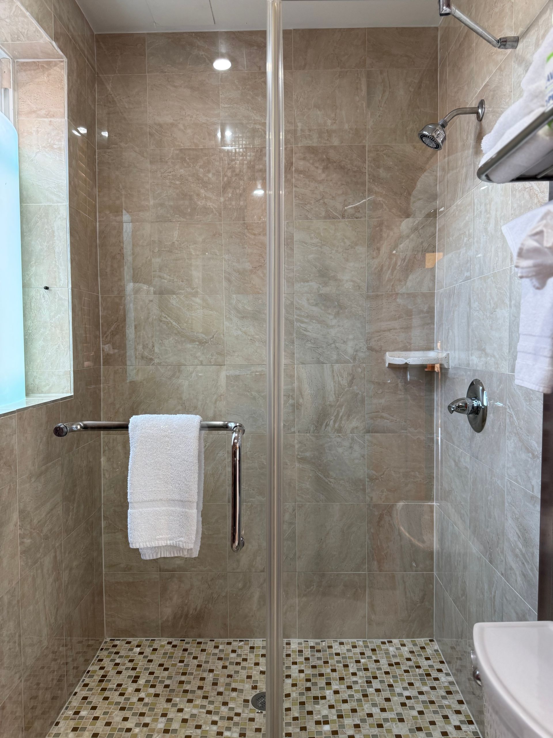 Shower with glass door, beige tile walls, pebble floor, and chrome fixtures. White towel hangs on the handle.