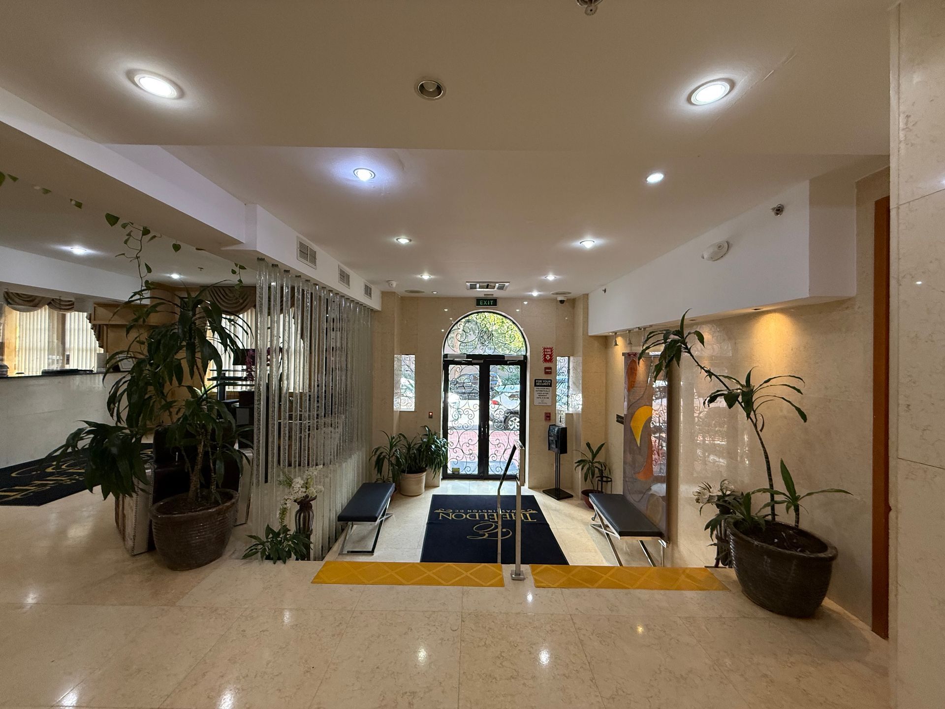 Lobby entrance with plants, benches, and a black doormat. Doors lead outside. Beige and white walls, recessed lighting.